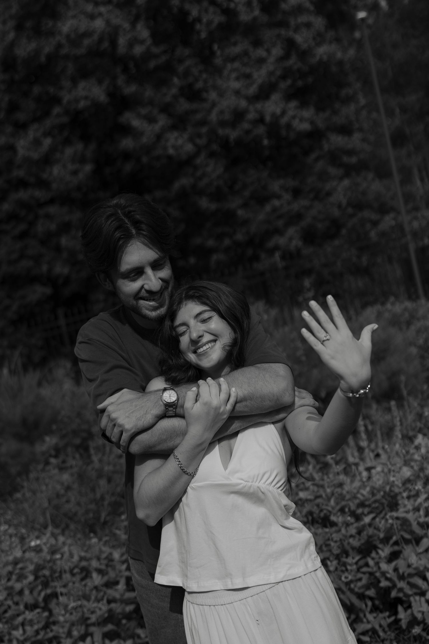 Black and white photo of couple hugging and laughing outdoors, woman showing off engagement ring.