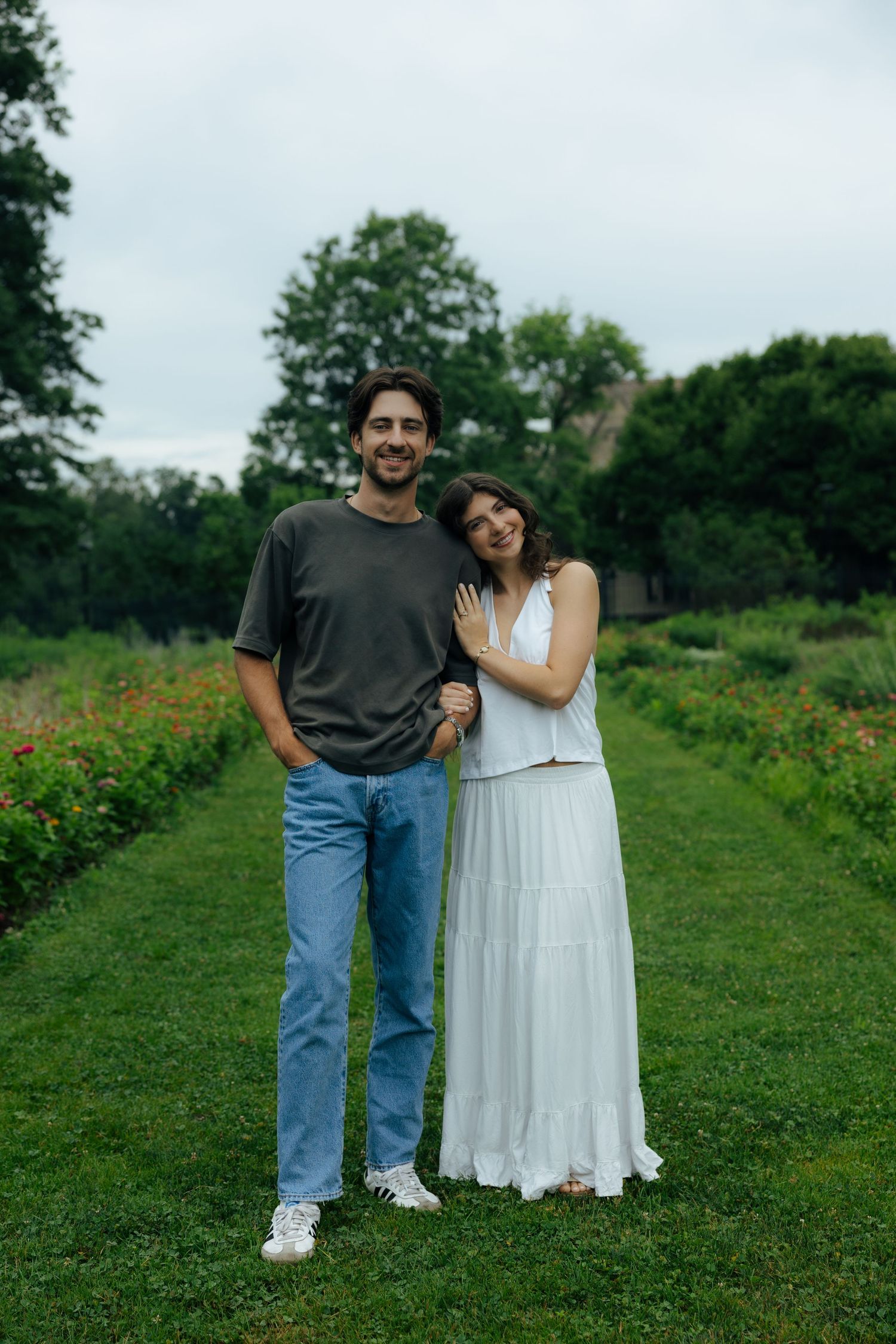 Happy couple posing in a lush garden path, woman in white dress leaning on man in gray tee and jeans.