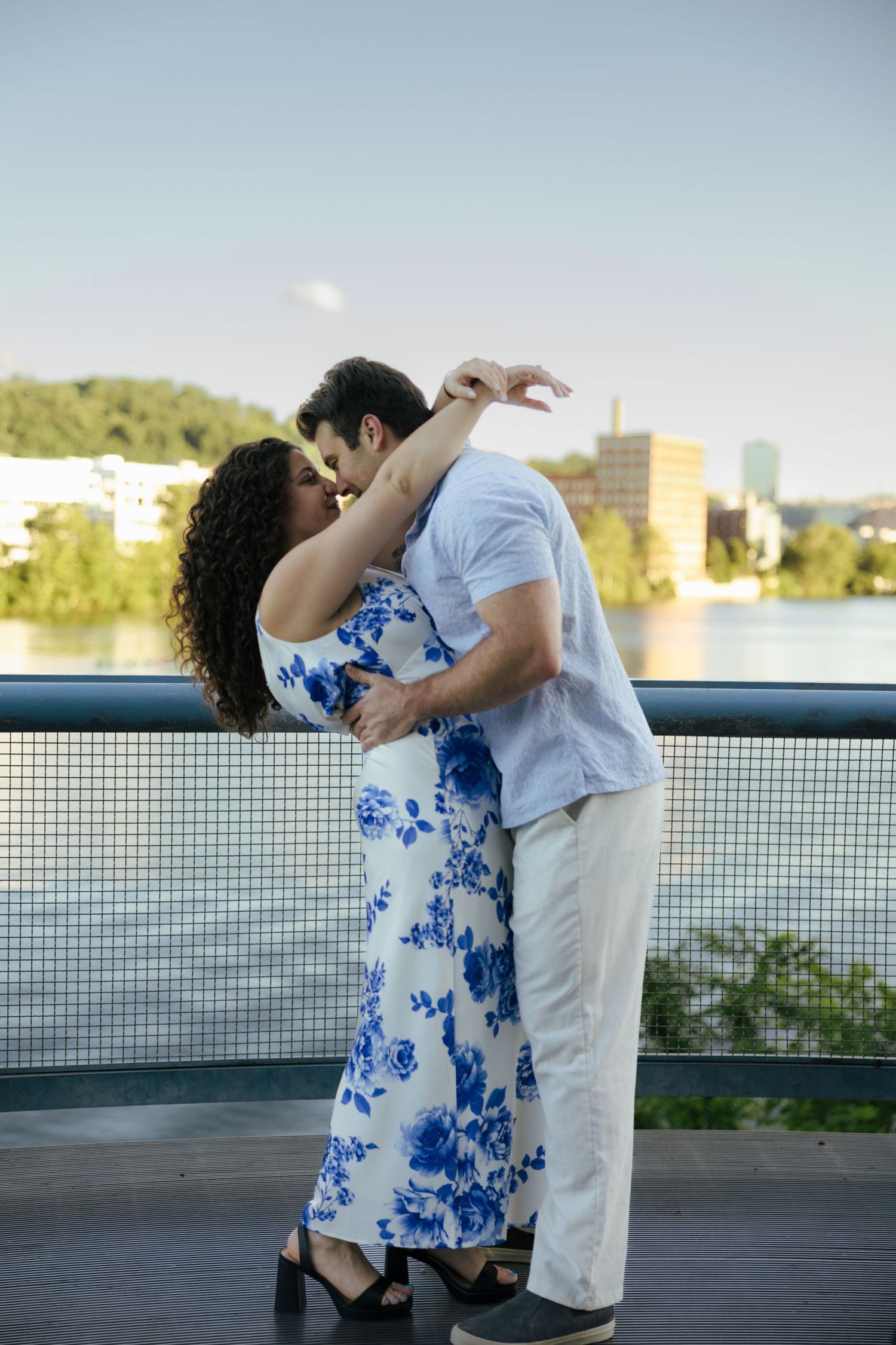 Engaged couple kissing on waterfront bridge, woman in white blue floral dress with city skyline behind.