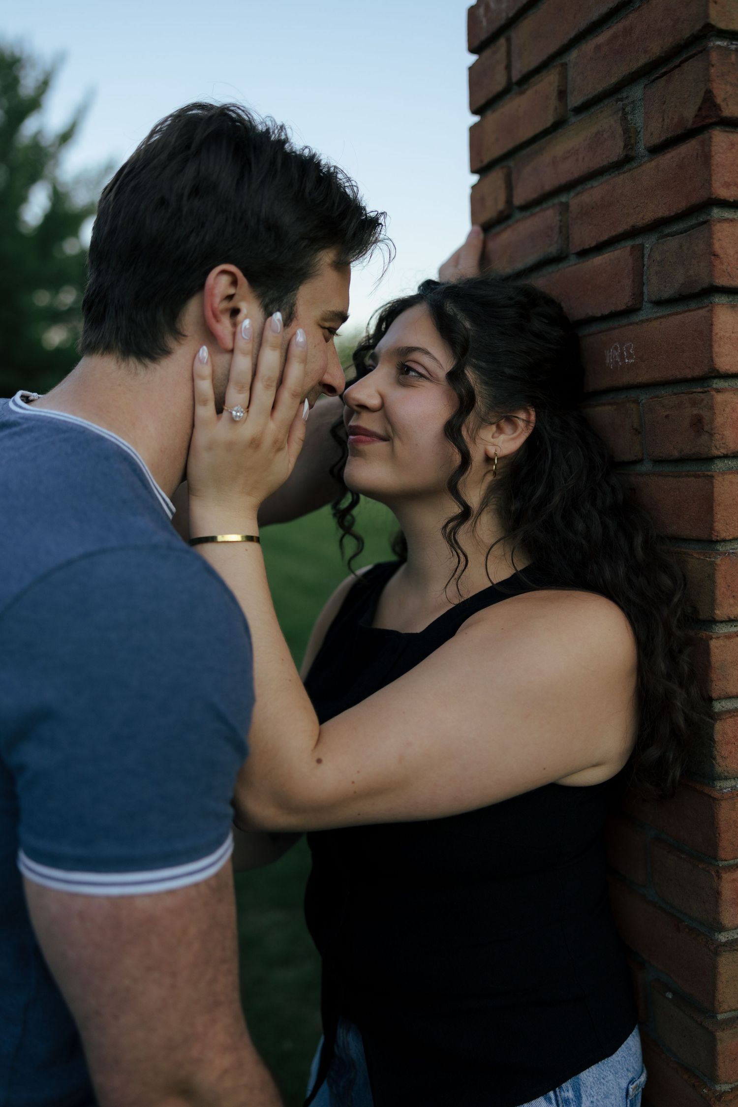 A young couple shares a romantic moment near a brick wall, with the woman tenderly holding the man's face close to hers.