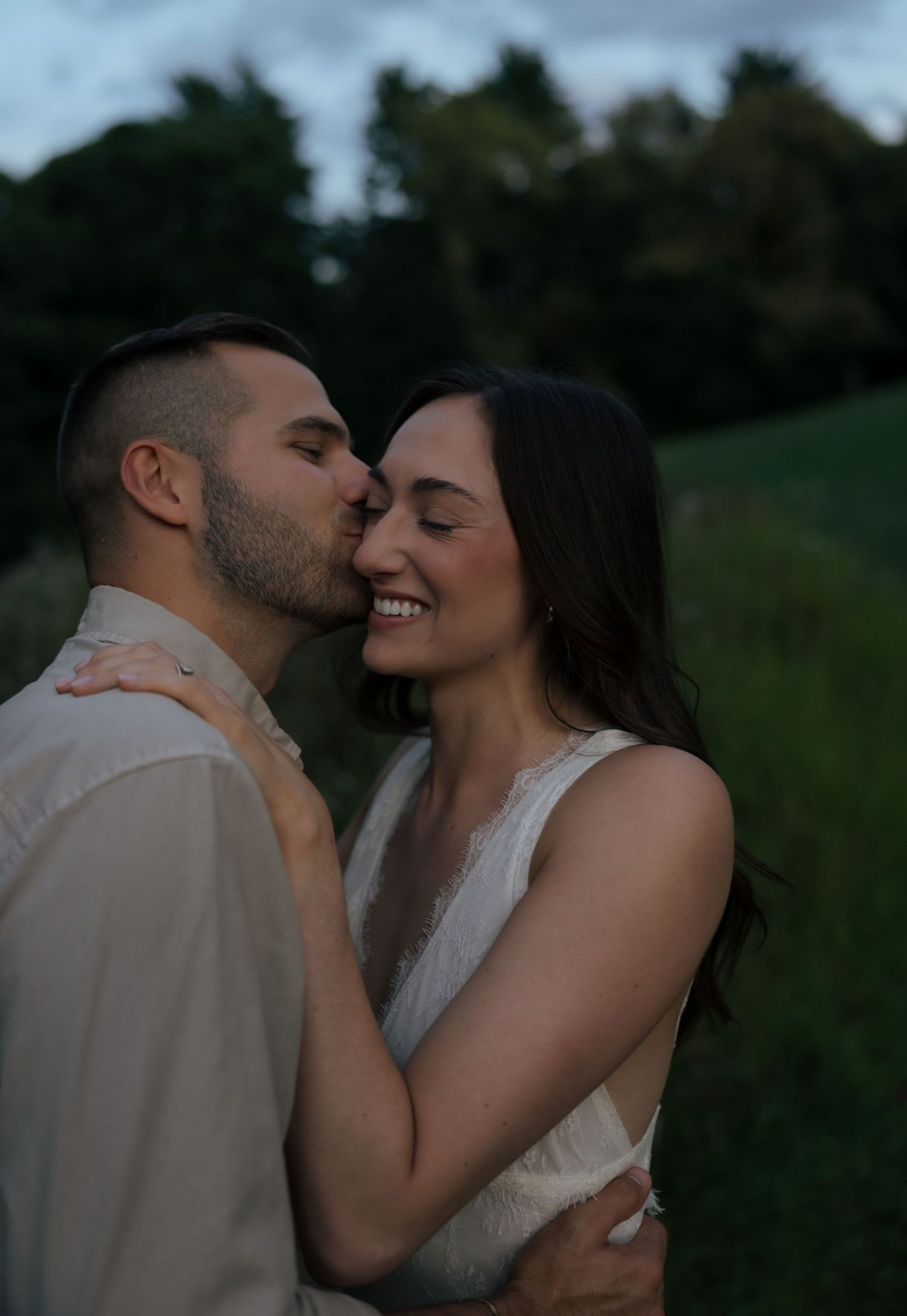 A couple shares a tender kiss at dusk, the woman in white smiling joyfully, surrounded by lush greenery.