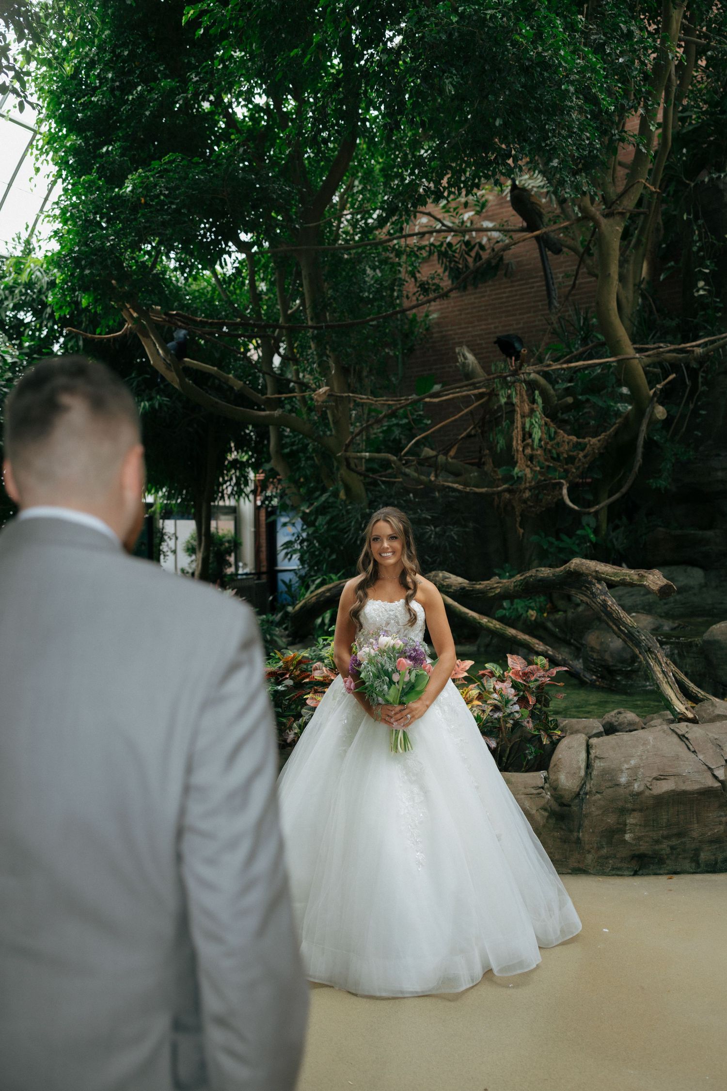 Smiling bride in a ball gown holding a bouquet during a first look at a lush indoor venue.