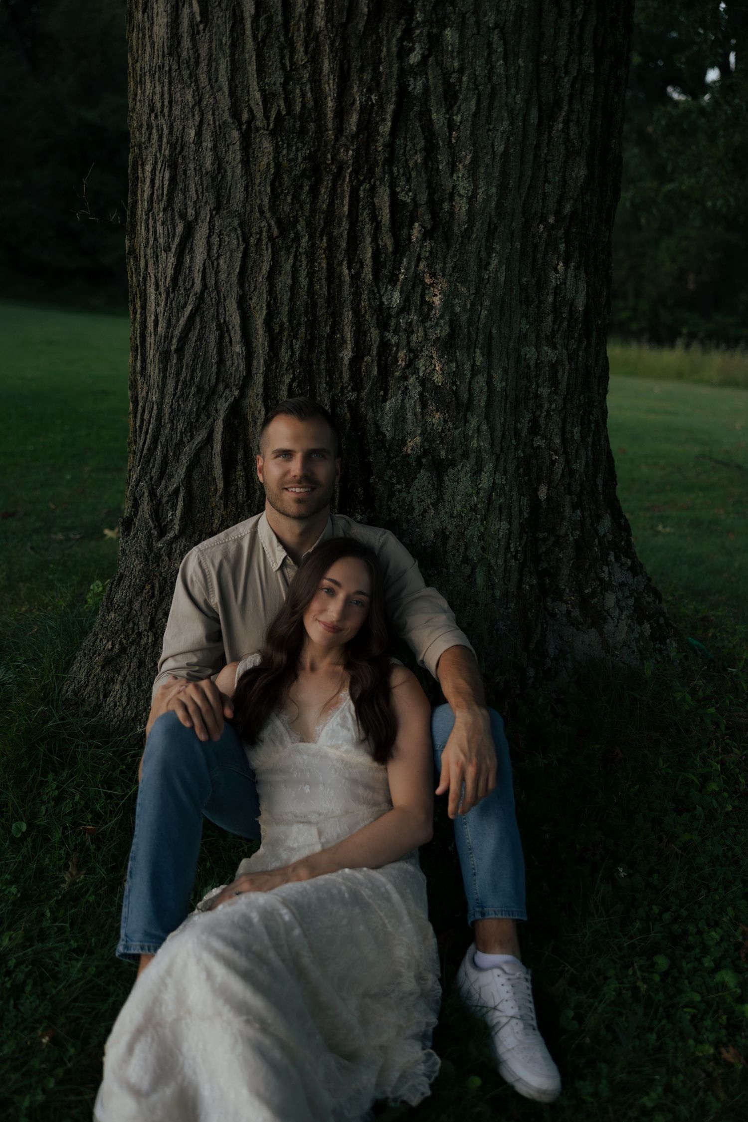 Bride and groom sit together leaning against massive tree trunk in evening park setting.
