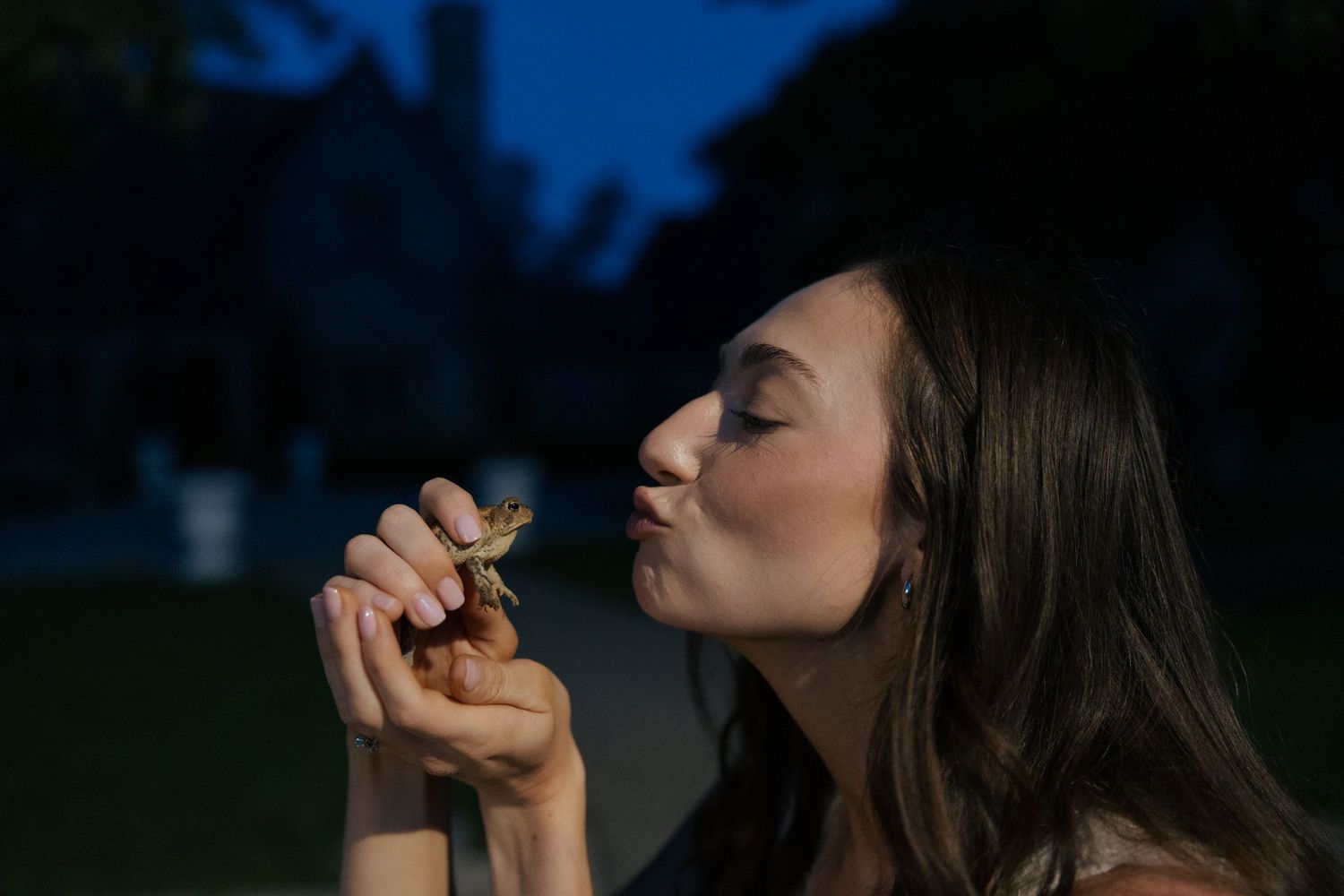 Young woman kissing a small frog she holds in her hands at twilight outdoors.