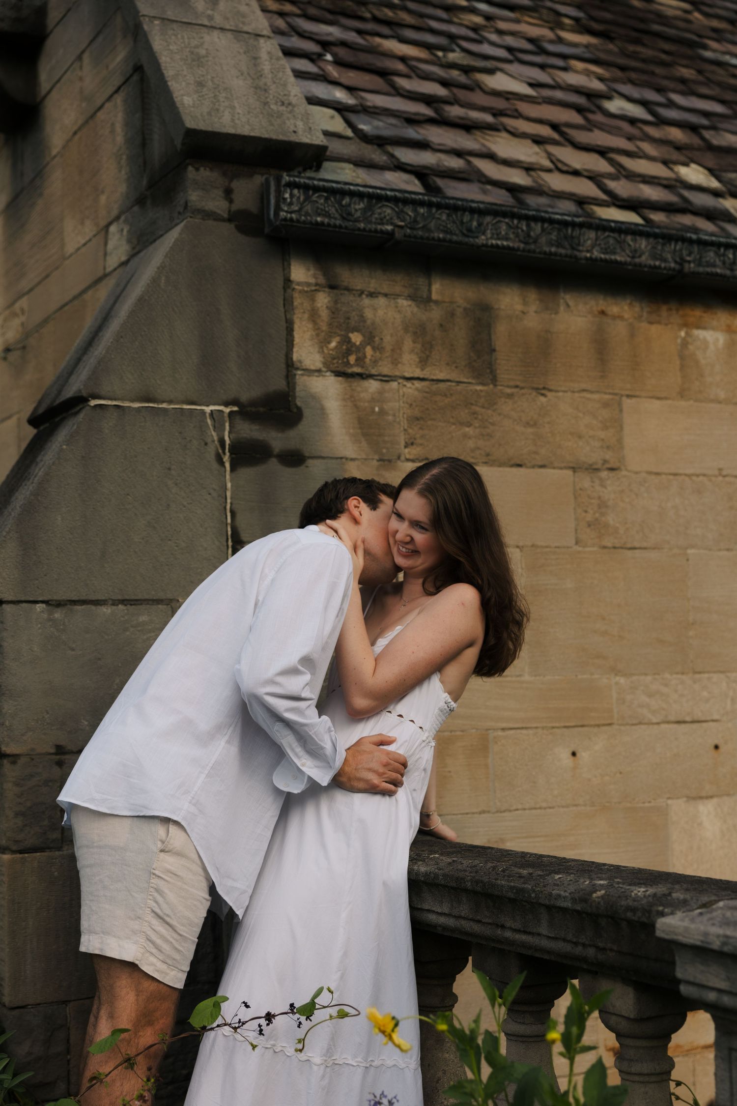 Couple sharing a tender moment against a historic stone wall, woman smiling in white dress.