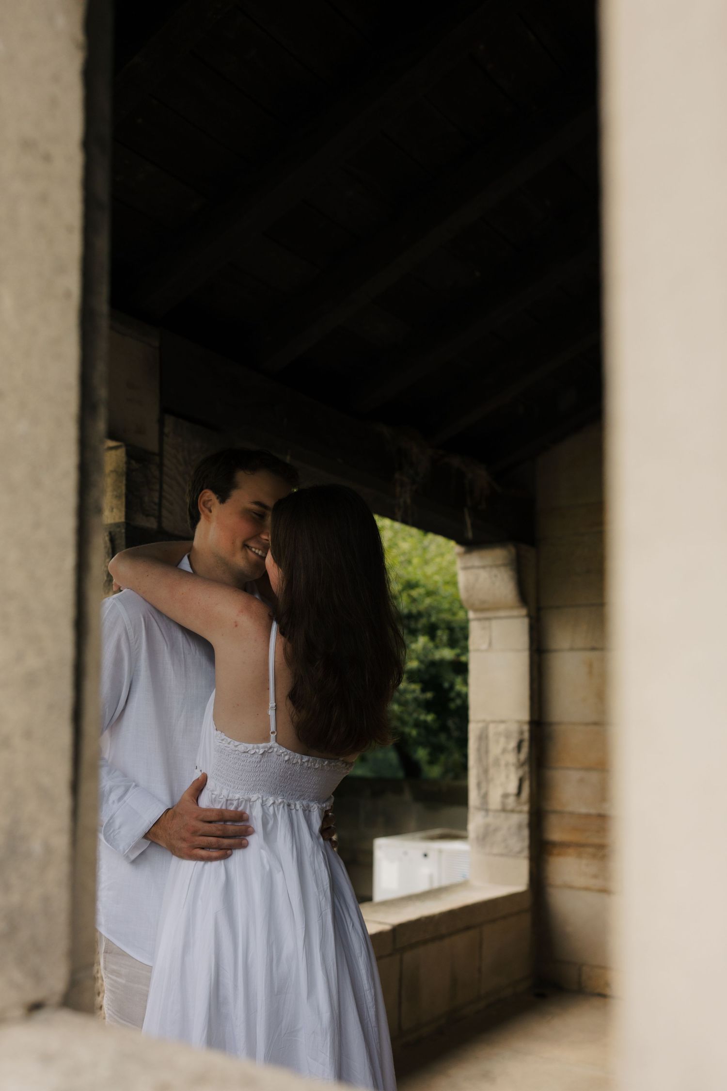 Couple embracing and smiling in a stone archway, woman in white dress with lace back, man in light blue shirt.