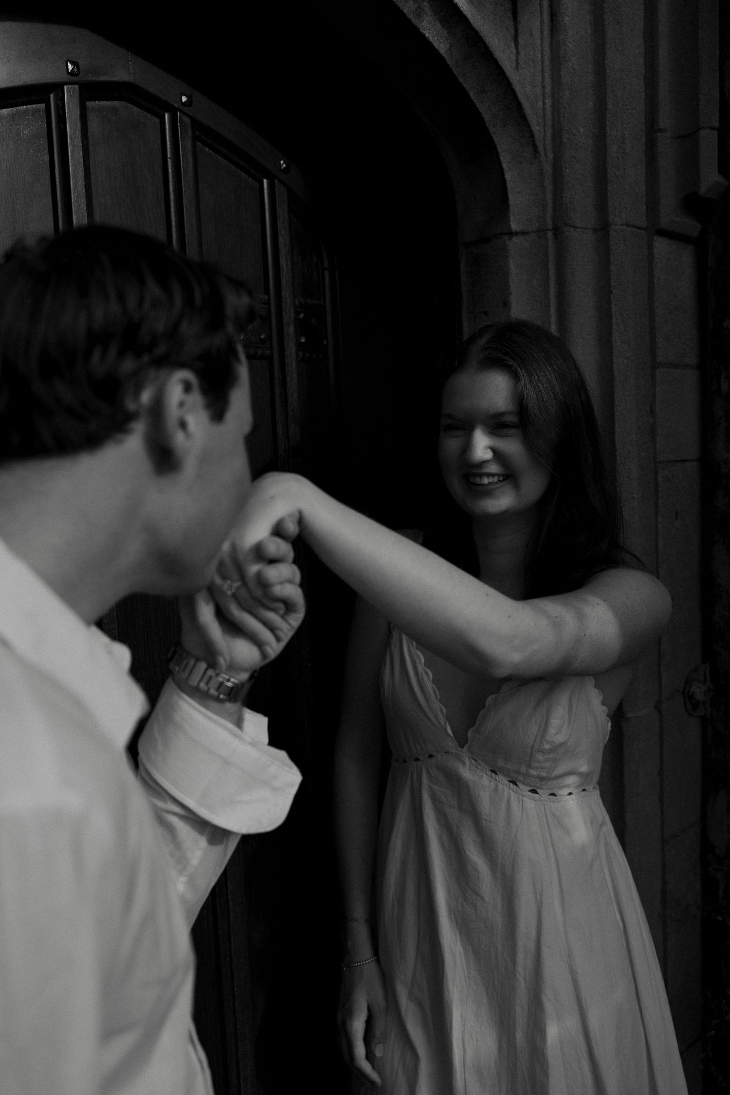 Black and white photo of a couple holding hands at an ornate stone doorway, woman smiling warmly.