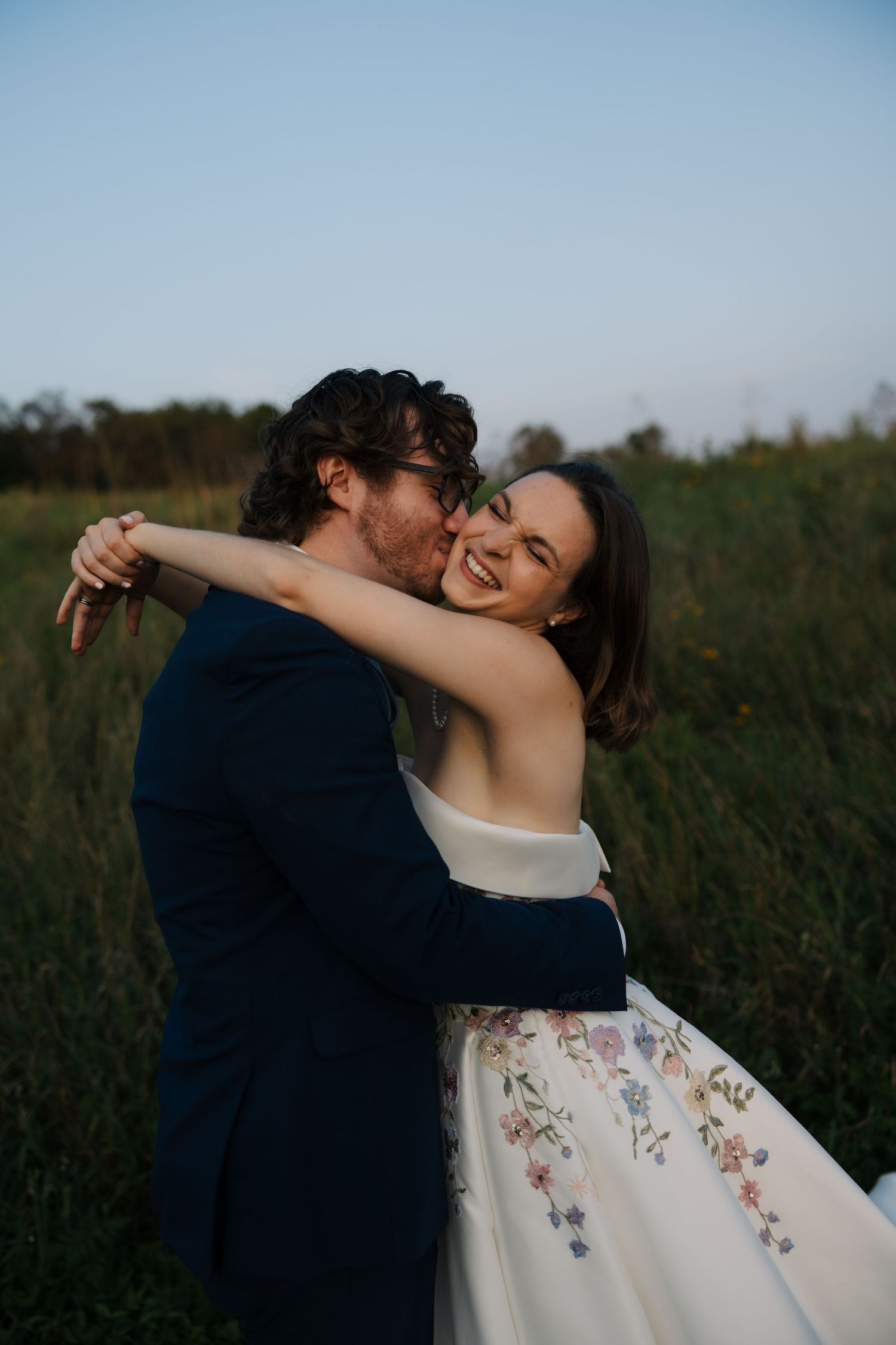 Groom kissing bride on the cheek as she smiles joyfully, both embracing in a lush green field at golden hour.