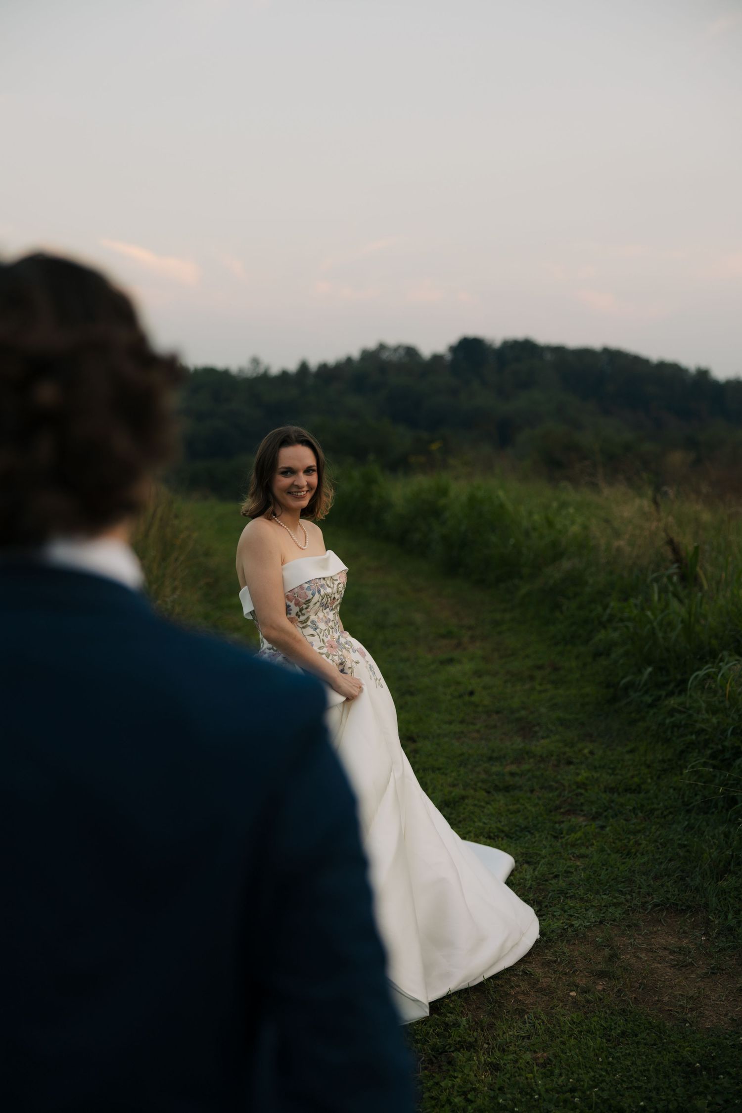 Bride in floral embroidered white gown smiles at groom on a lush green countryside path at dusk.