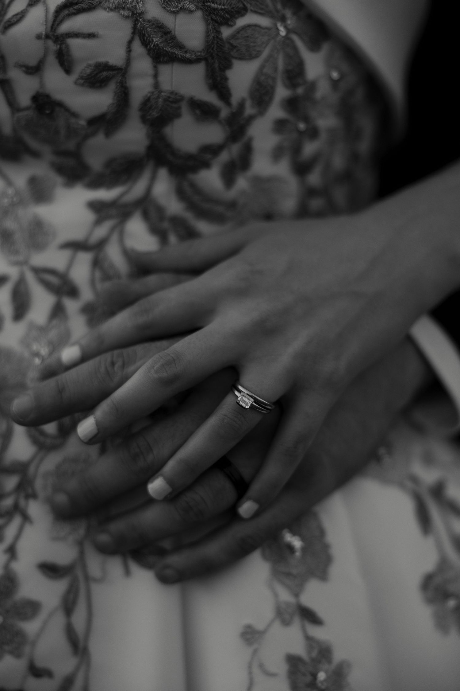 Close-up black and white photo of bride and groom's hands showing wedding ring on floral lace gown.