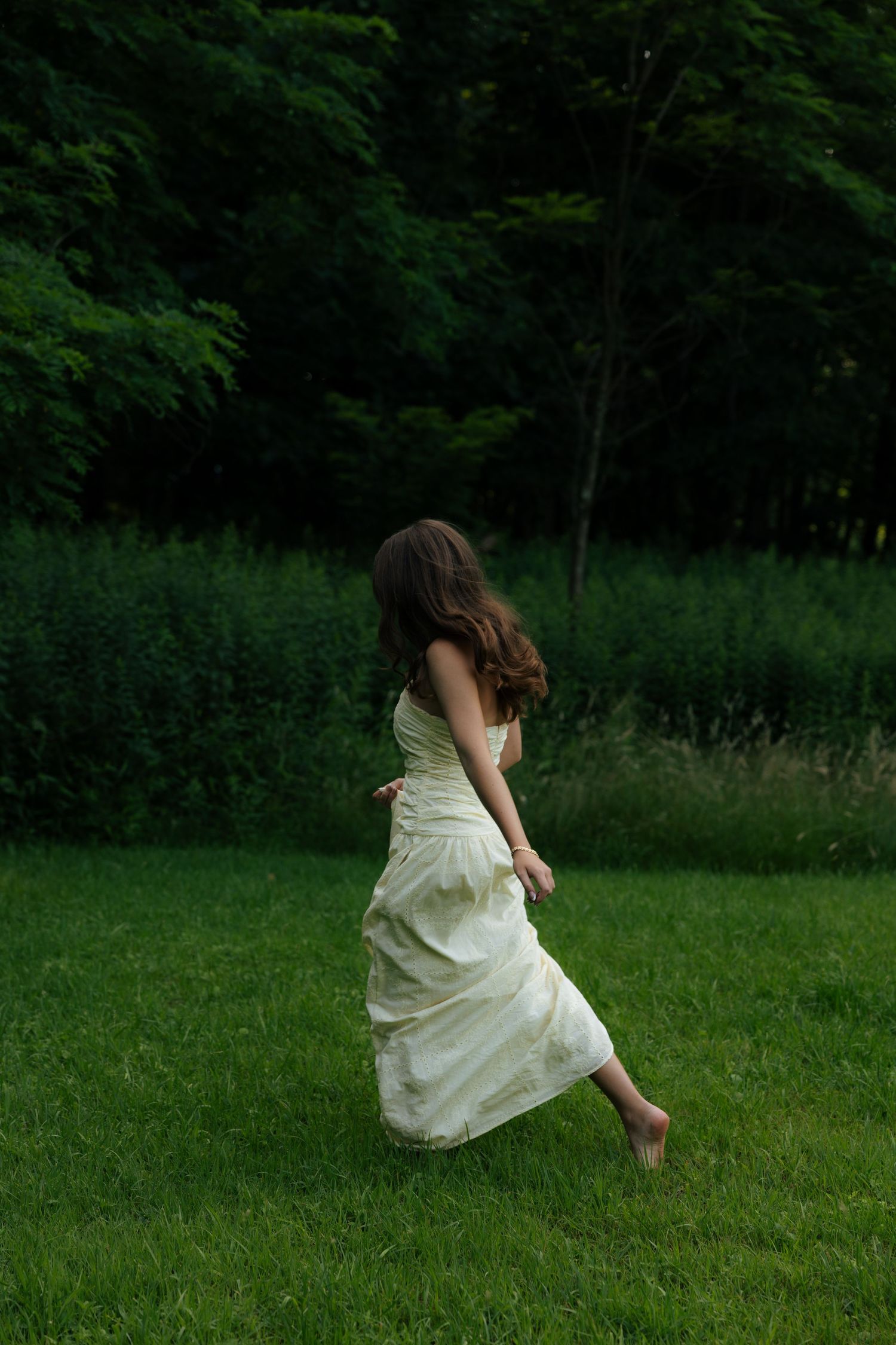 A barefoot woman in a flowing white dress runs across a lush green lawn toward a dark forest.