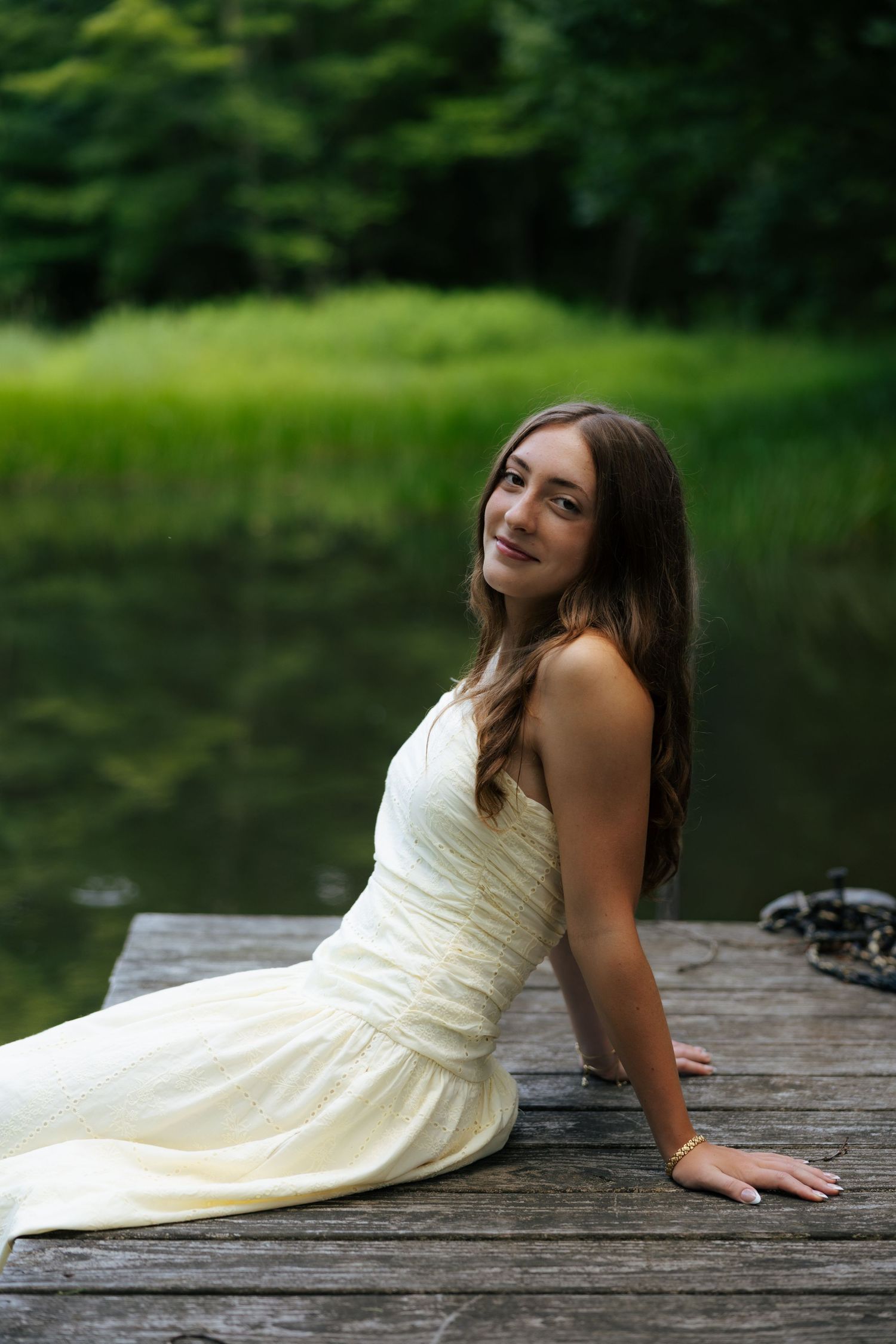 Young woman in white dress sitting gracefully on wooden dock beside calm reflective lake.