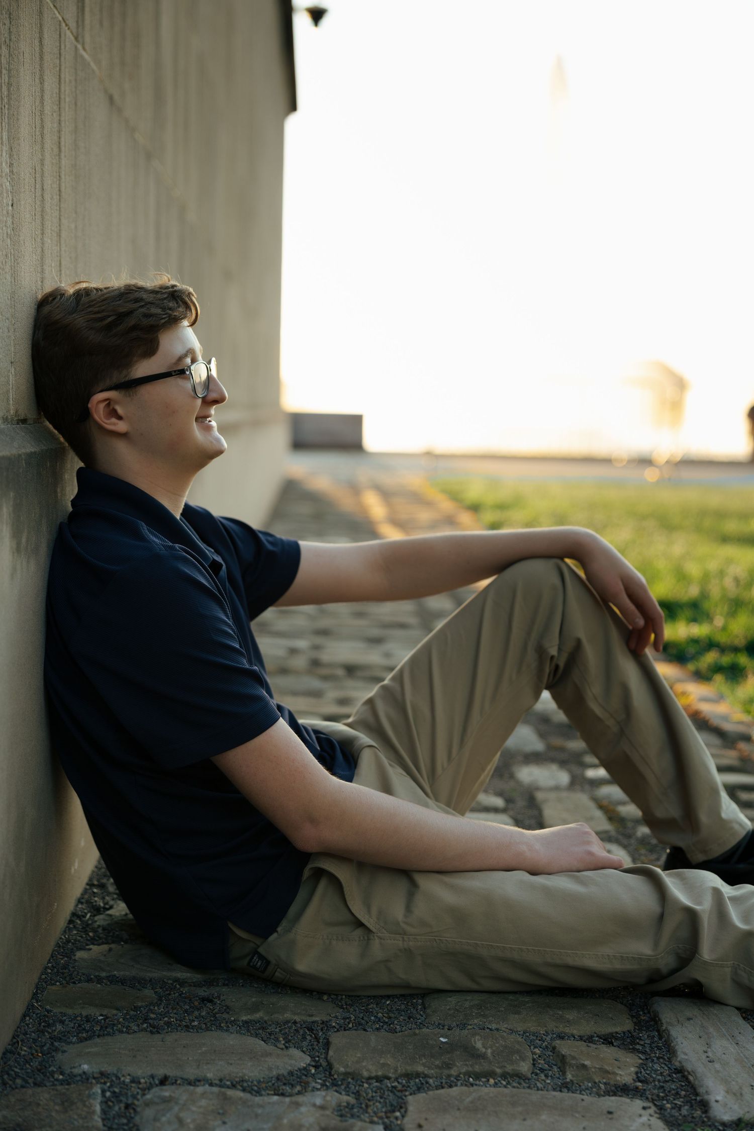 Teenage boy in navy polo sitting against a wall on cobblestone ground smiling at sunset.