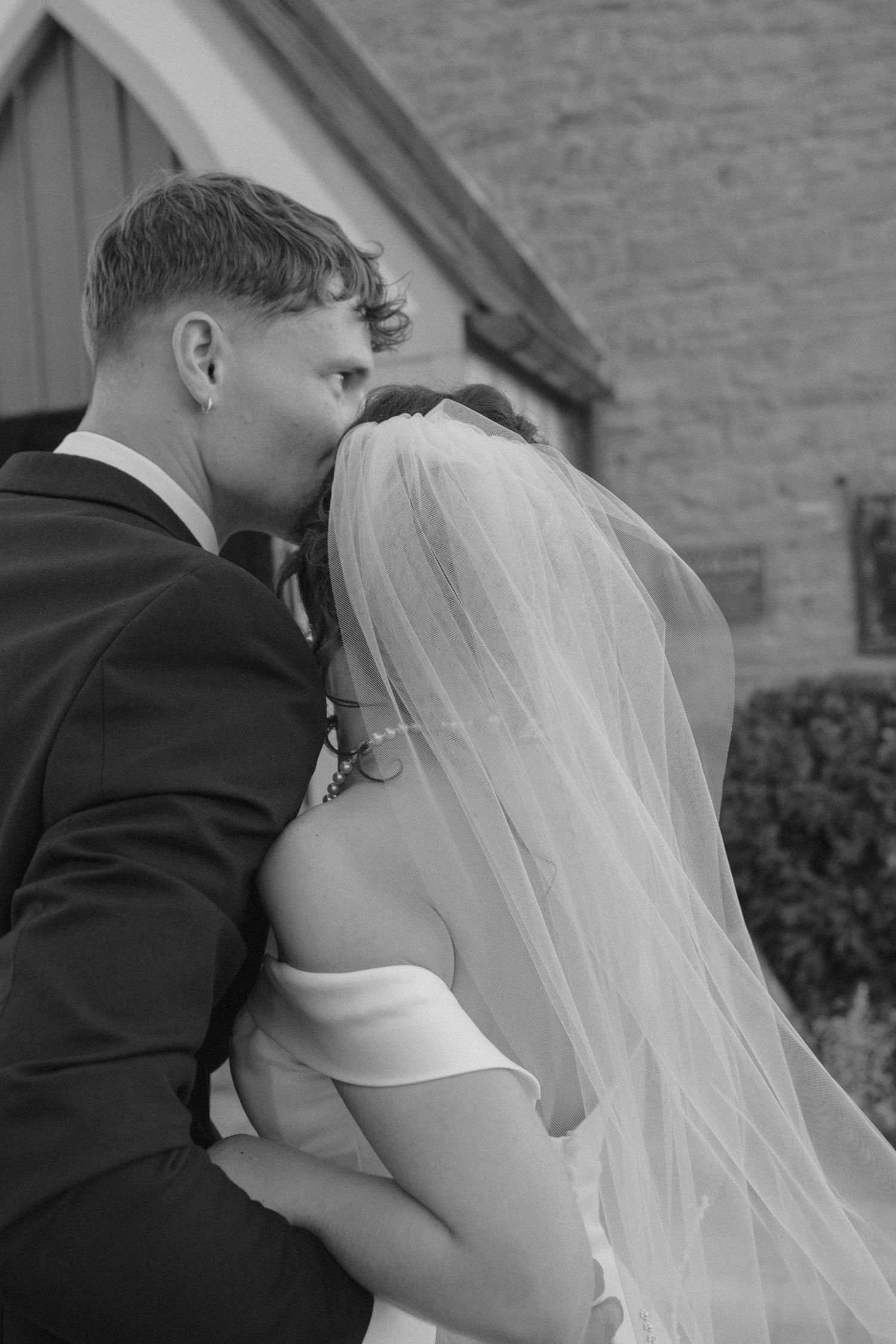 Black and white photo of a groom kissing his bride on the forehead outside a church on their wedding day.