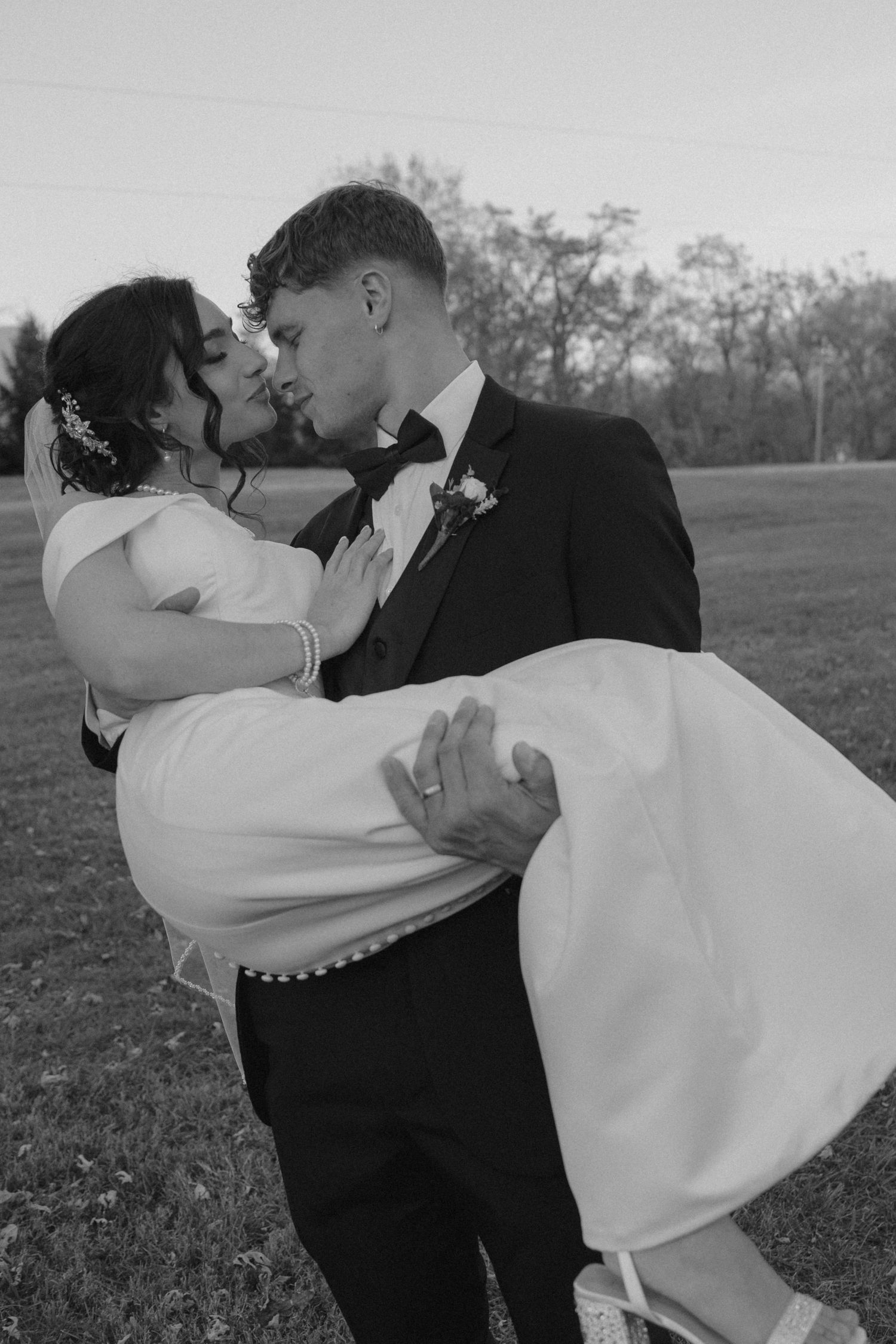 Groom carries bride in his arms as they share a kiss in an open field during their wedding celebration.