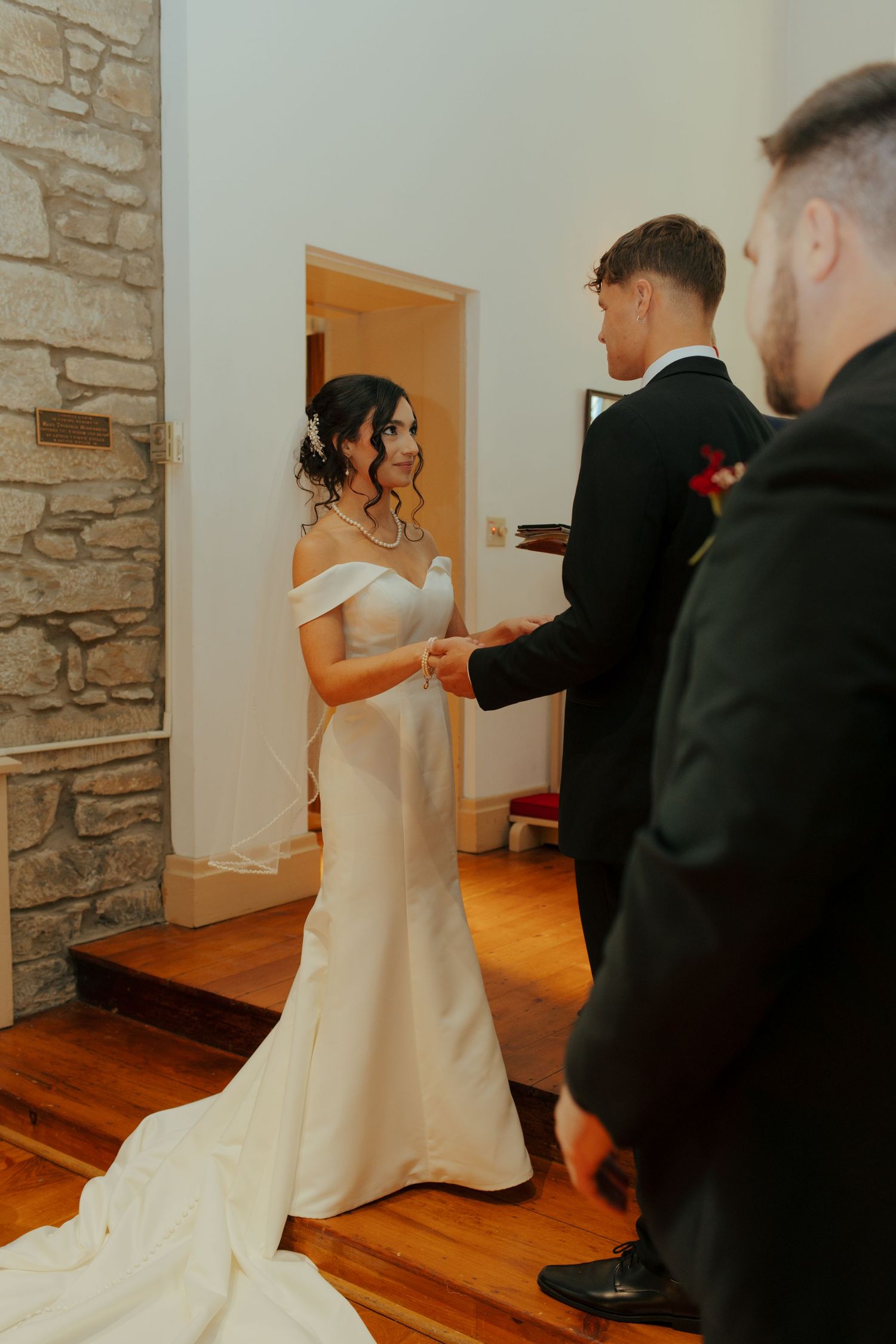 Bride in off-shoulder white gown holds hands with groom during intimate indoor wedding ceremony at stone church.
