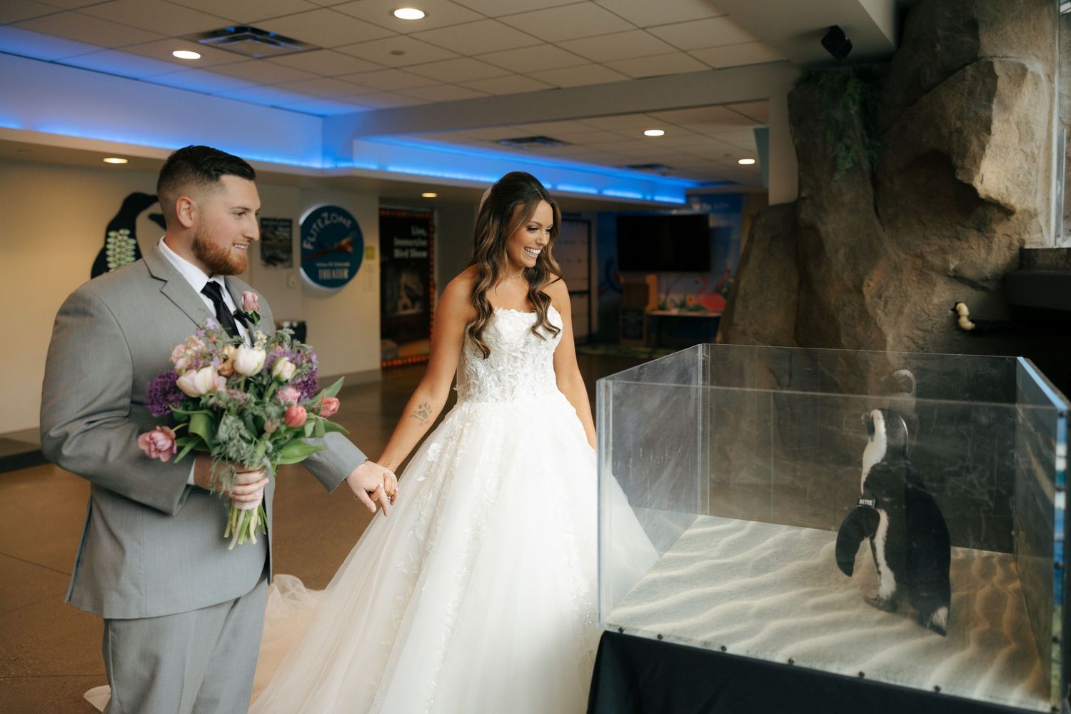 Bride and groom in wedding attire holding flowers beside a penguin exhibit at an aquarium venue.