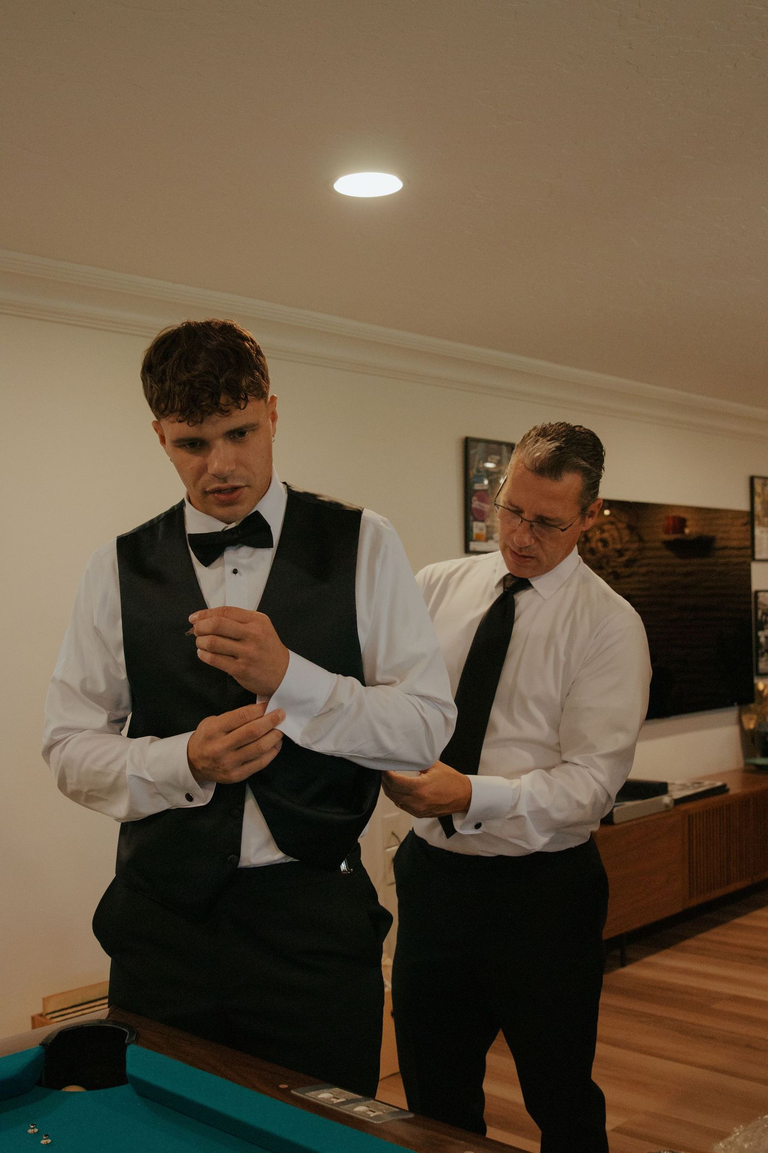 Father helps groom with cufflinks while getting ready in room with pool table before wedding.