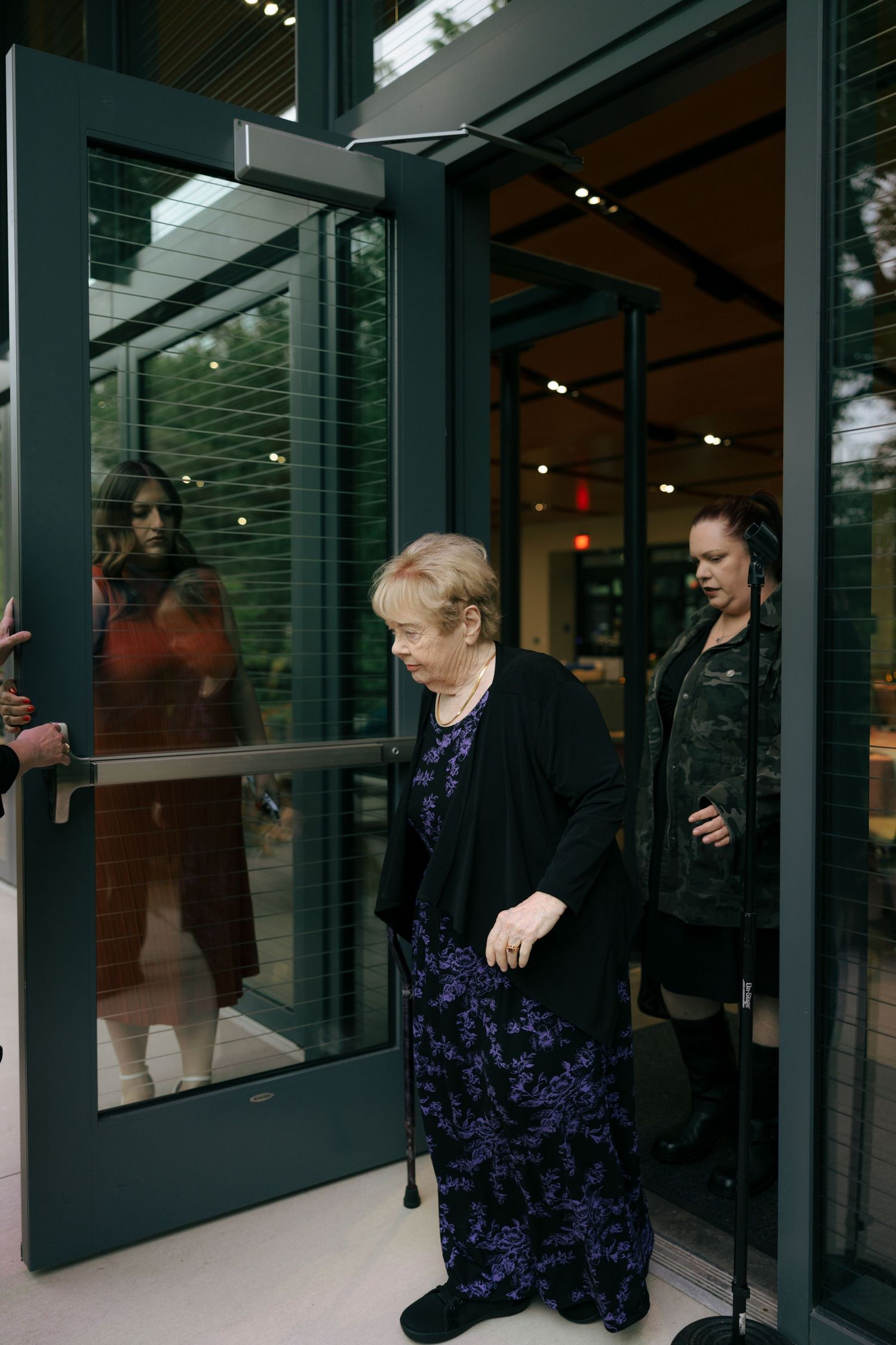 Elderly woman in blue floral dress exits glass door building, assisted by woman in dark outfit standing behind her.