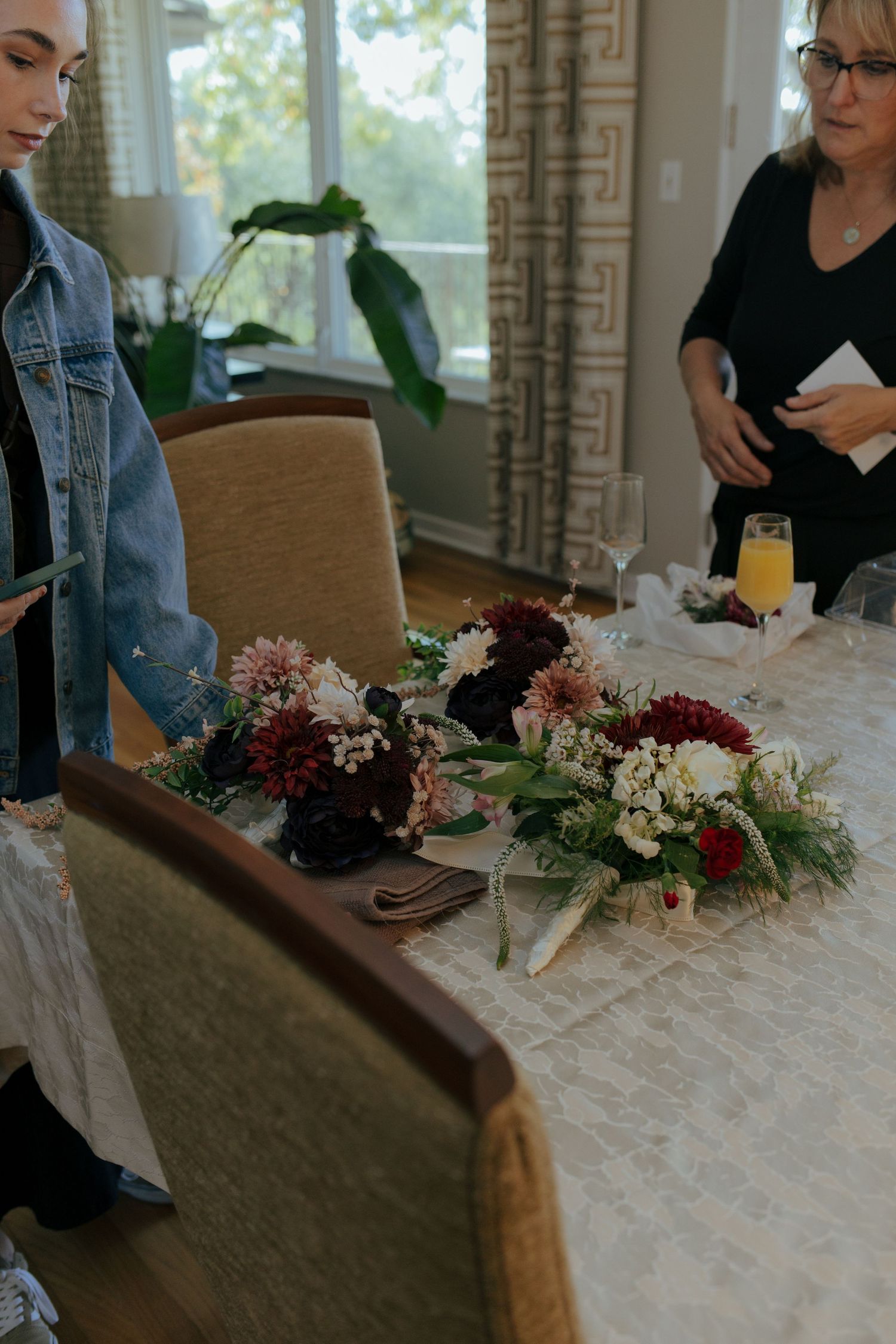 Two women arranging elegant floral centerpieces with burgundy and white flowers on a dining table.