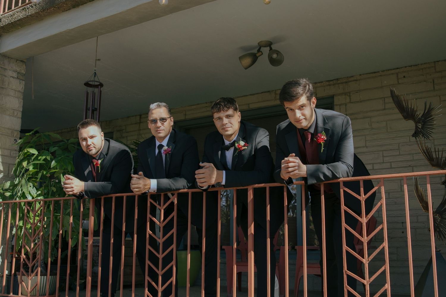 Four groomsmen in dark suits lean on a decorative iron railing on a porch, wearing boutonnieres for a wedding.