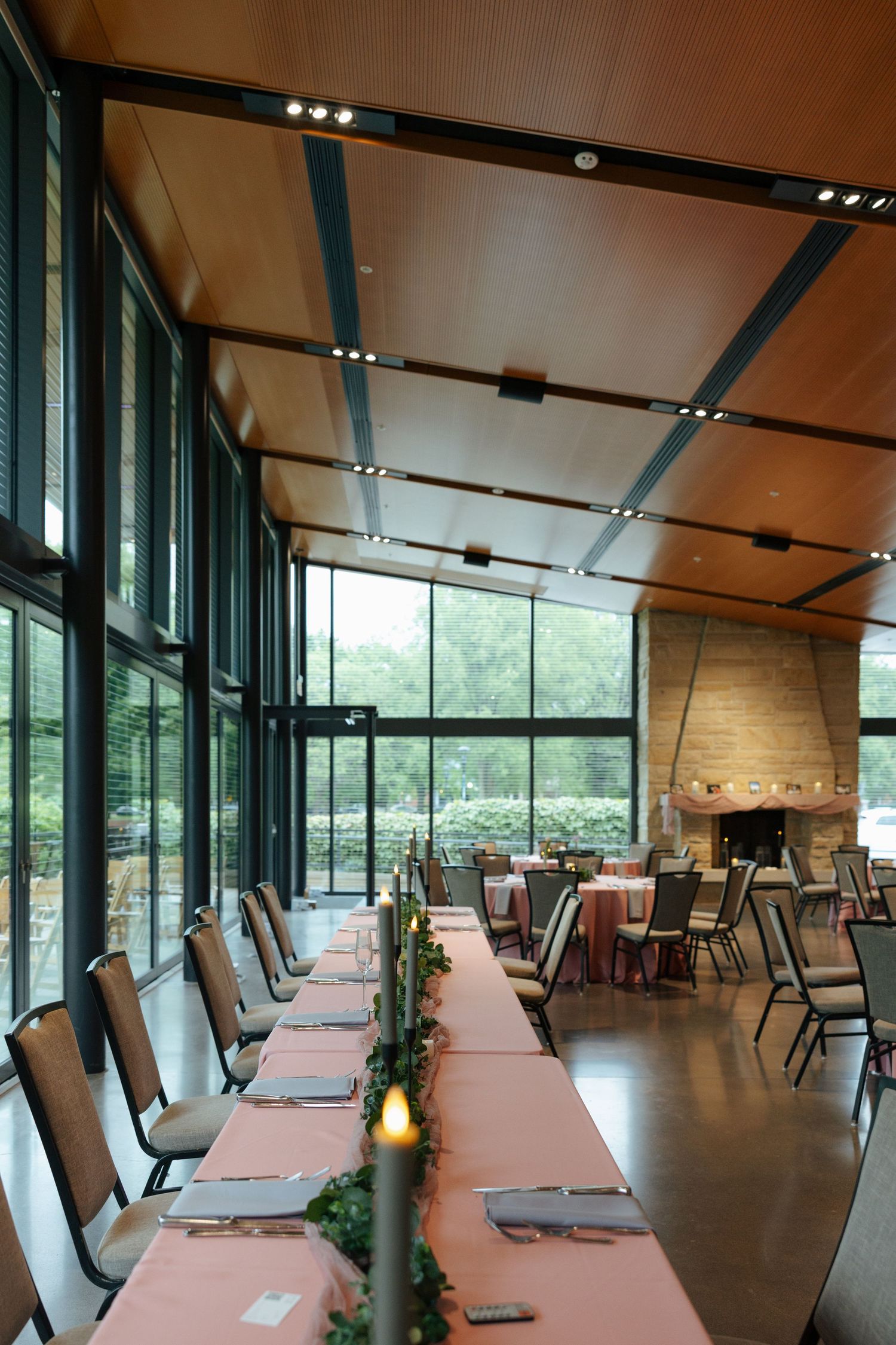 Elegant banquet hall with floor-to-ceiling windows, long pink-draped table, candles, and a stone fireplace in background.