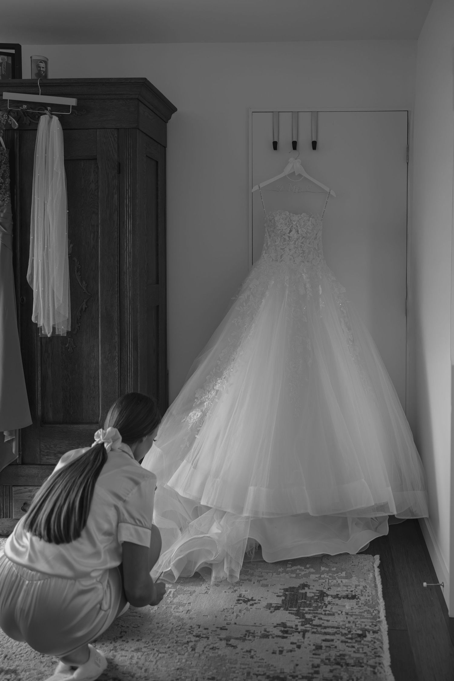 Black and white photo of bride crouching to arrange her ball gown wedding dress hanging on wall hooks.