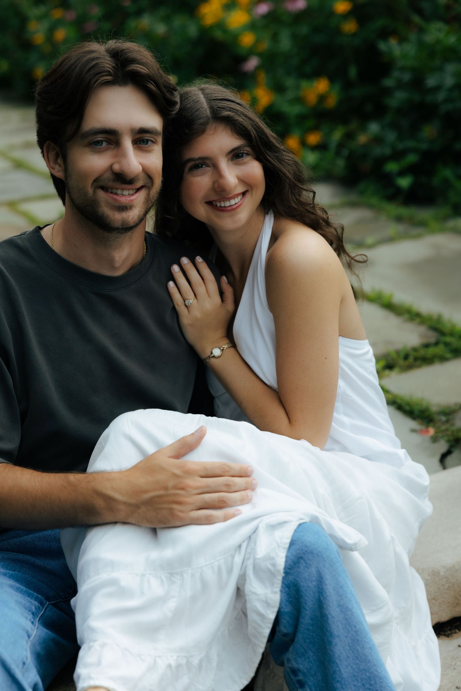Smiling couple sitting together outdoors in a garden, woman in white dress leaning on man in dark shirt wearing jeans.