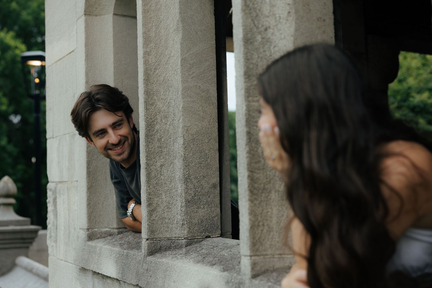 Smiling bearded man peers through stone columns at dark-haired woman during outdoor photo session.