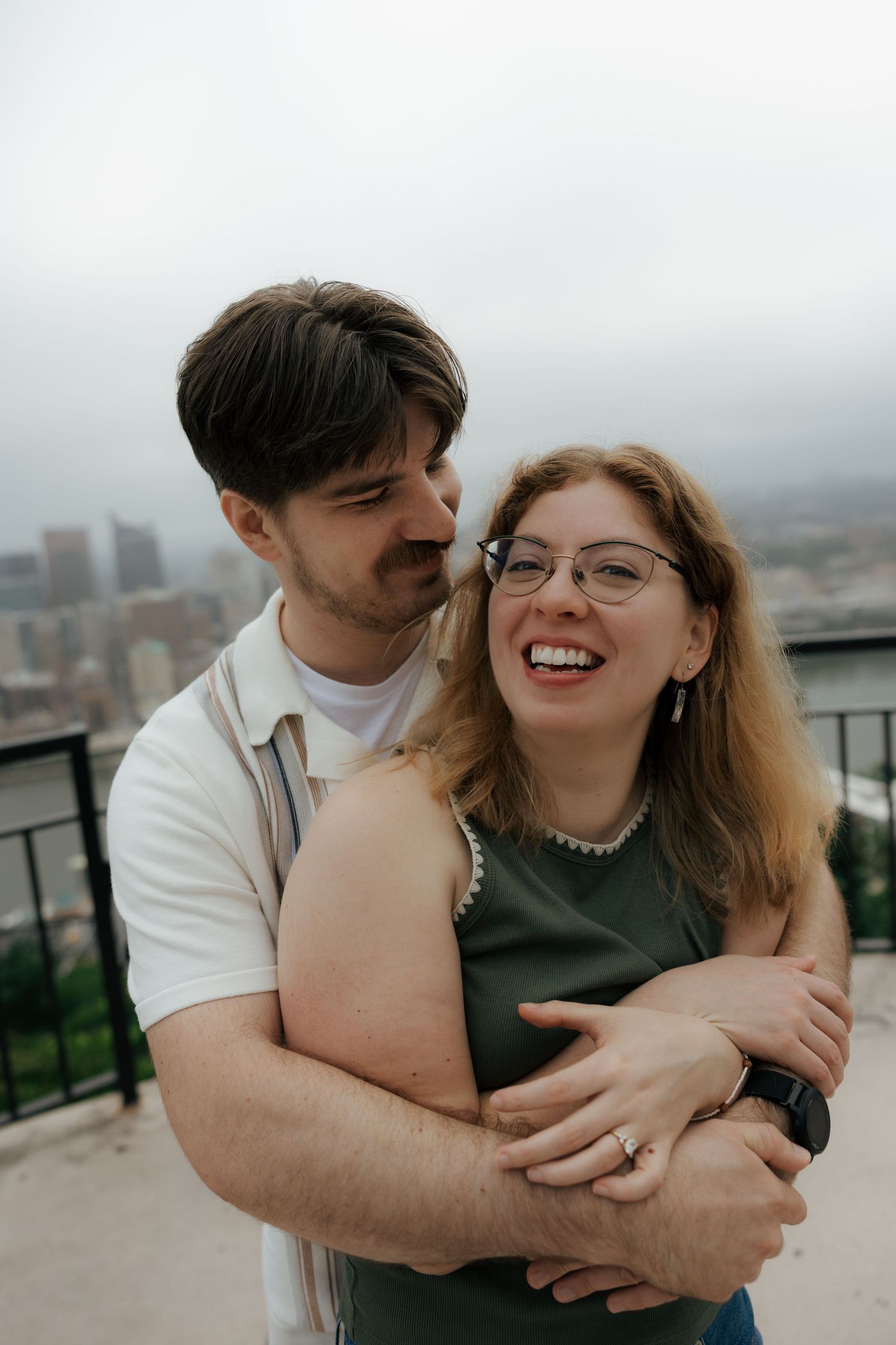 A joyful couple embraces on a rooftop overlook, woman laughing in green top and glasses, man hugging her from behind.