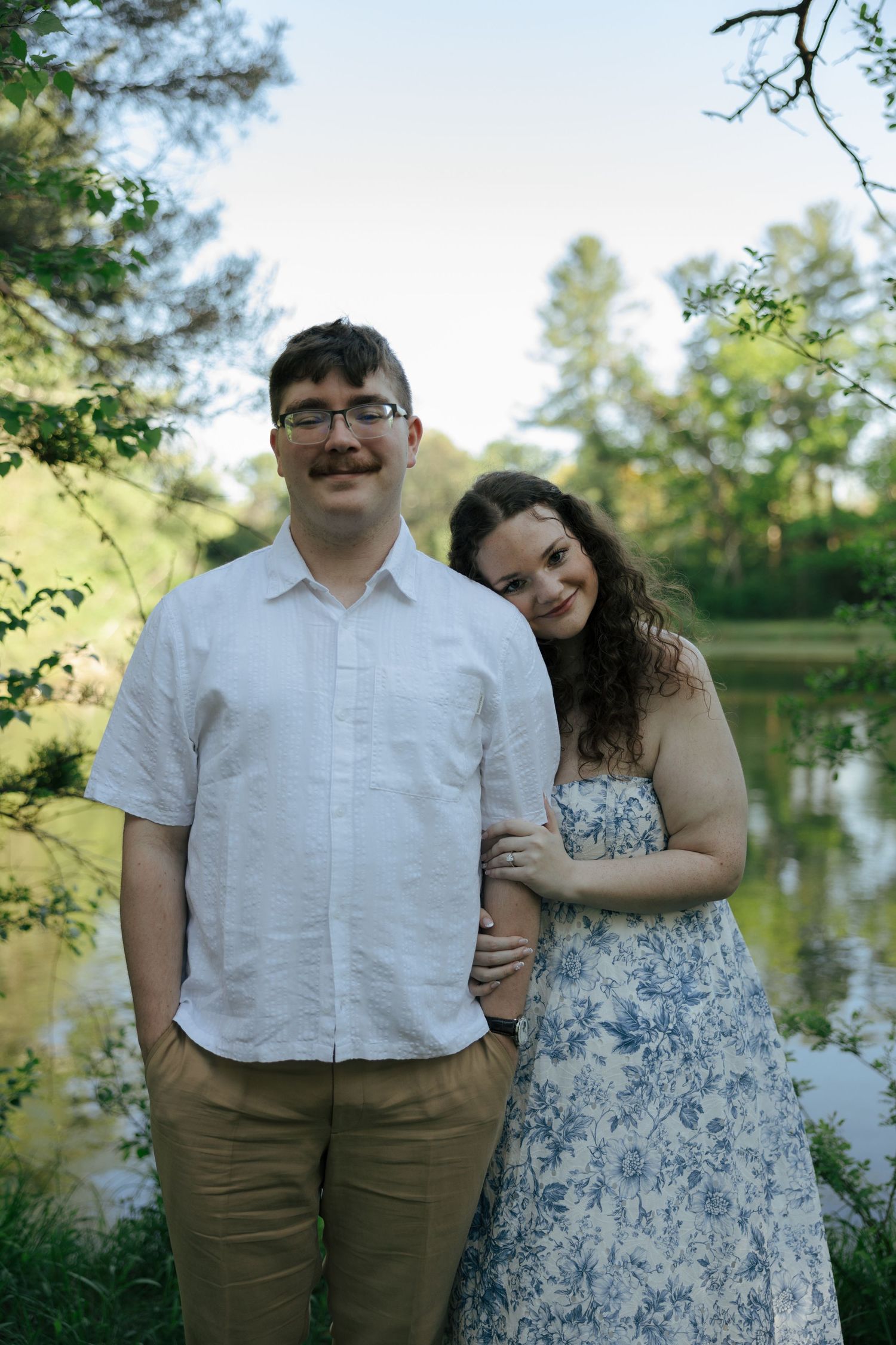 Smiling couple standing together near a reflective pond, woman in blue floral dress leaning on mans shoulder.