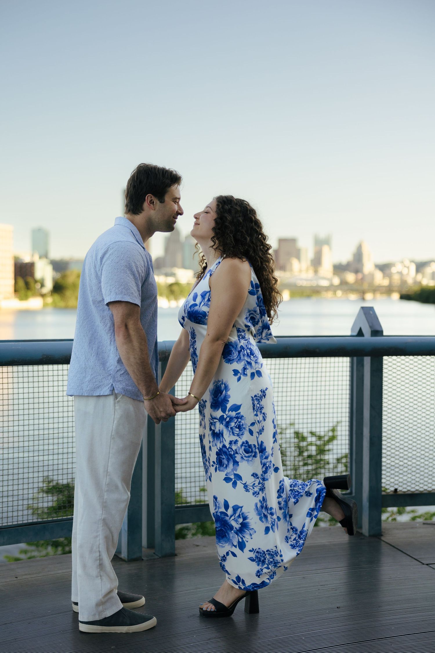 Couple holding hands facing each other on bridge with river and city skyline in background.