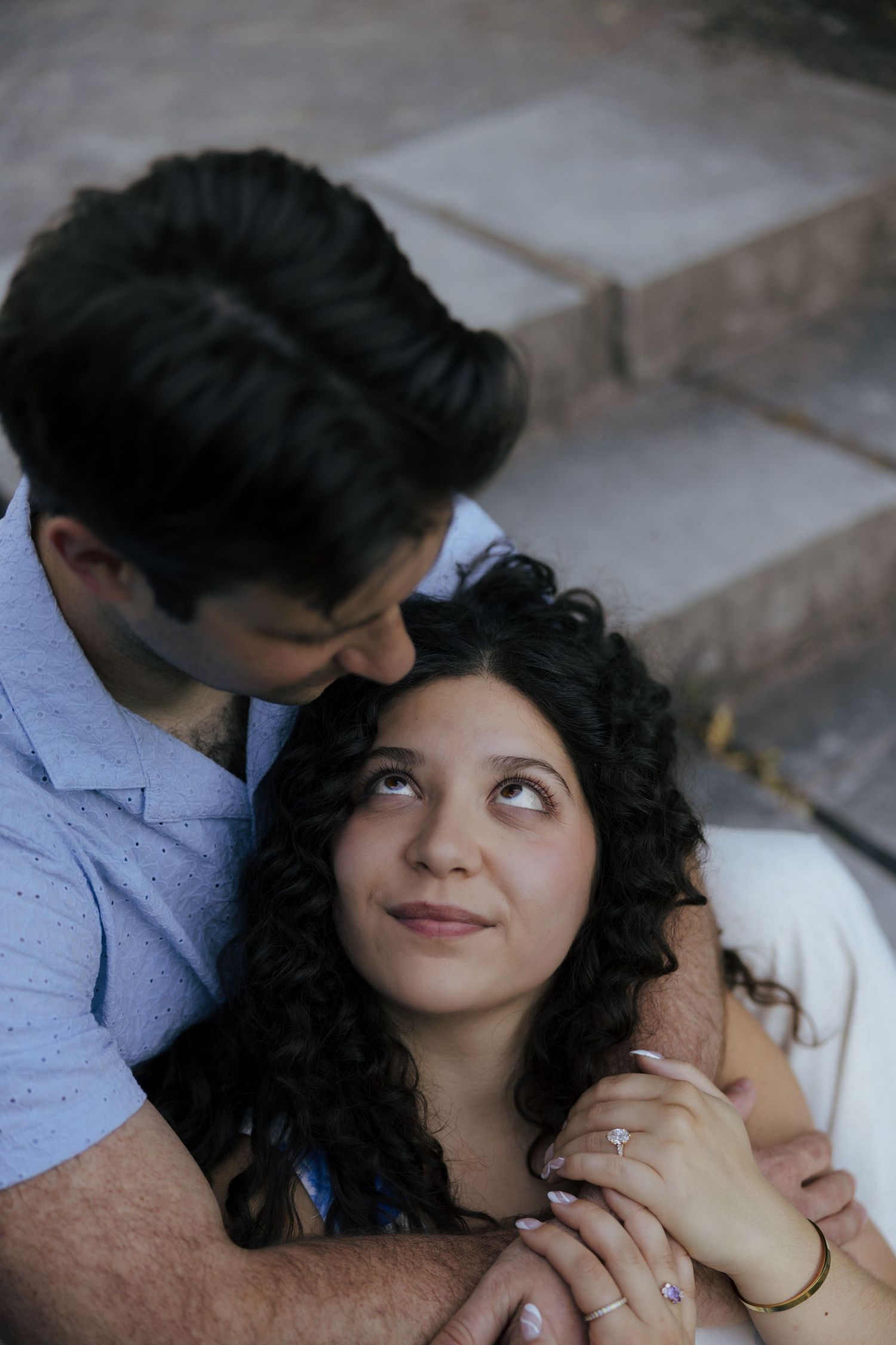 Couple sitting on steps, woman with curly hair looks up lovingly at man embracing her, engagement ring visible.