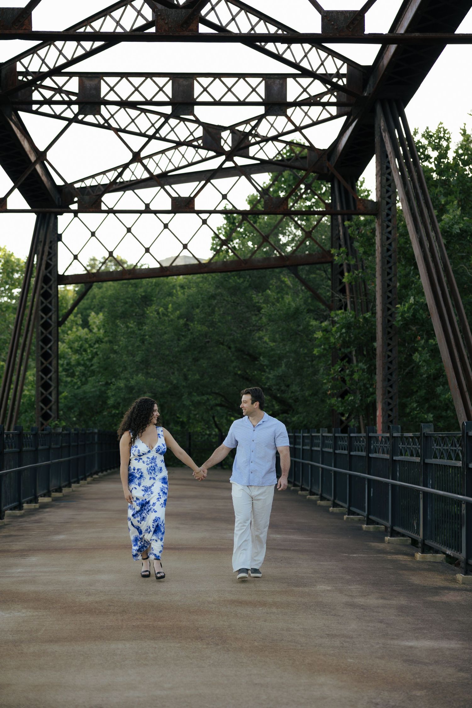 Couple holding hands walking across an iron bridge surrounded by green trees.