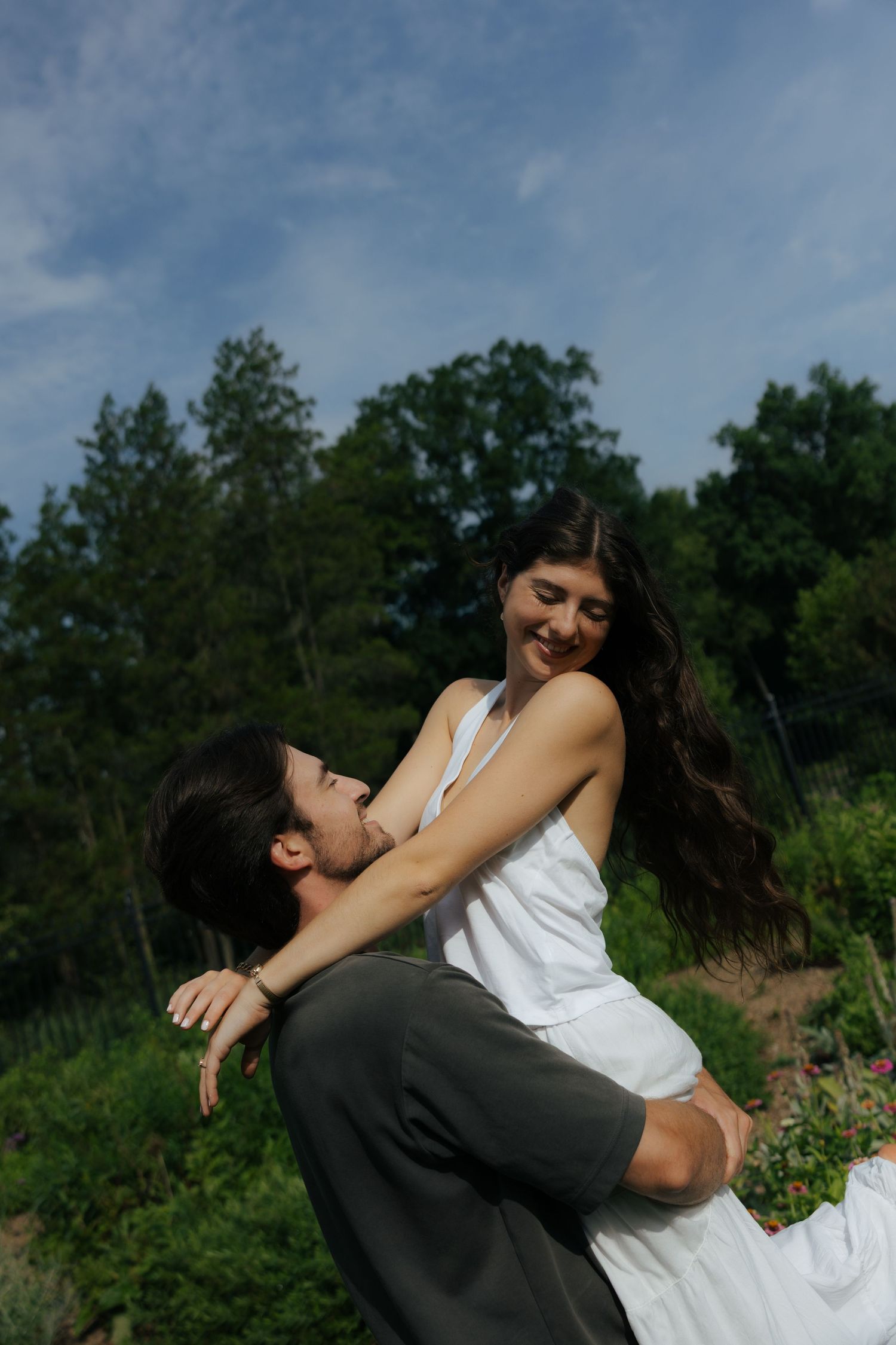 A man lifts a smiling woman in a white dress outdoors, surrounded by lush green trees under a partly cloudy sky.