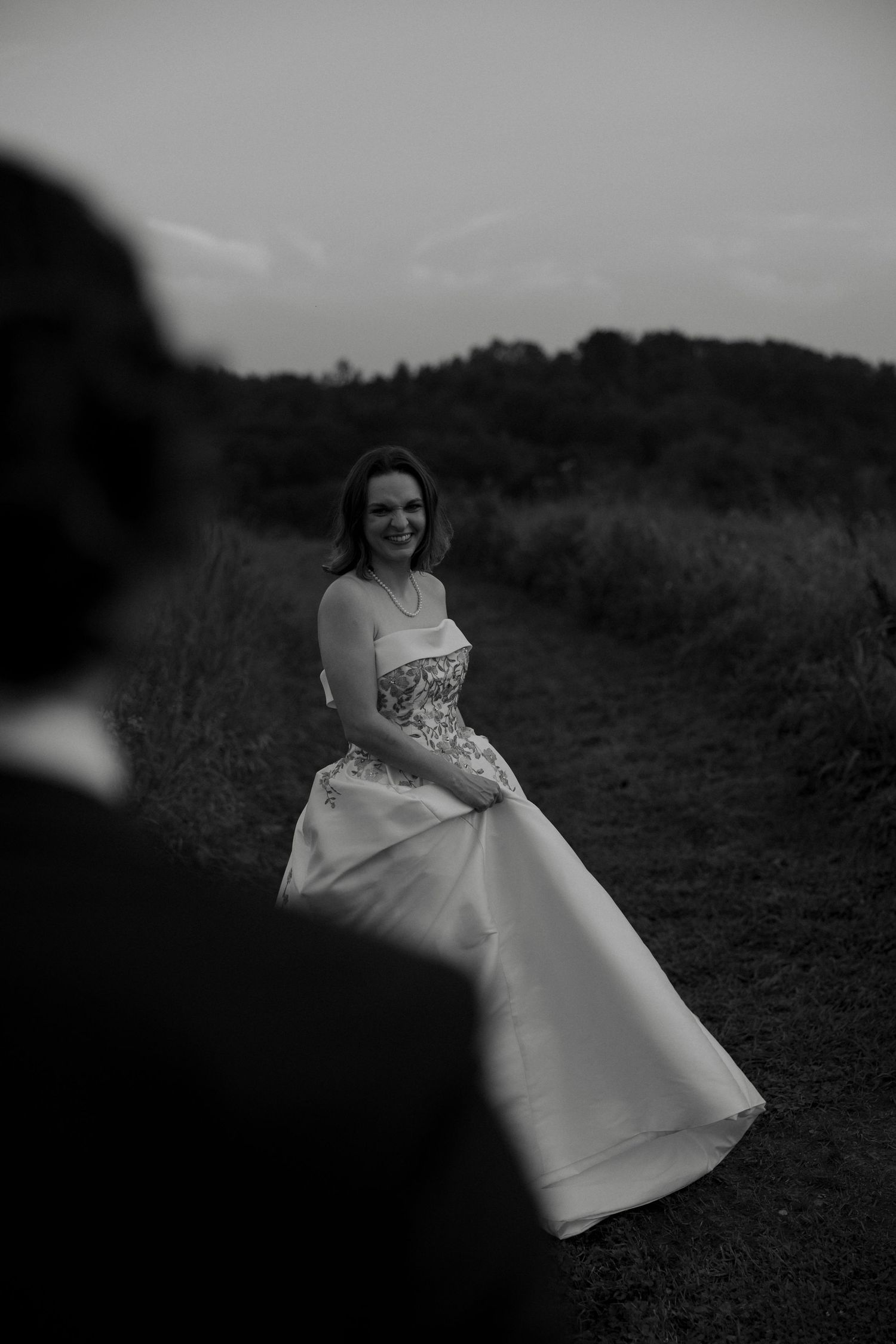 Black and white photo of smiling bride in strapless gown on outdoor path at dusk.