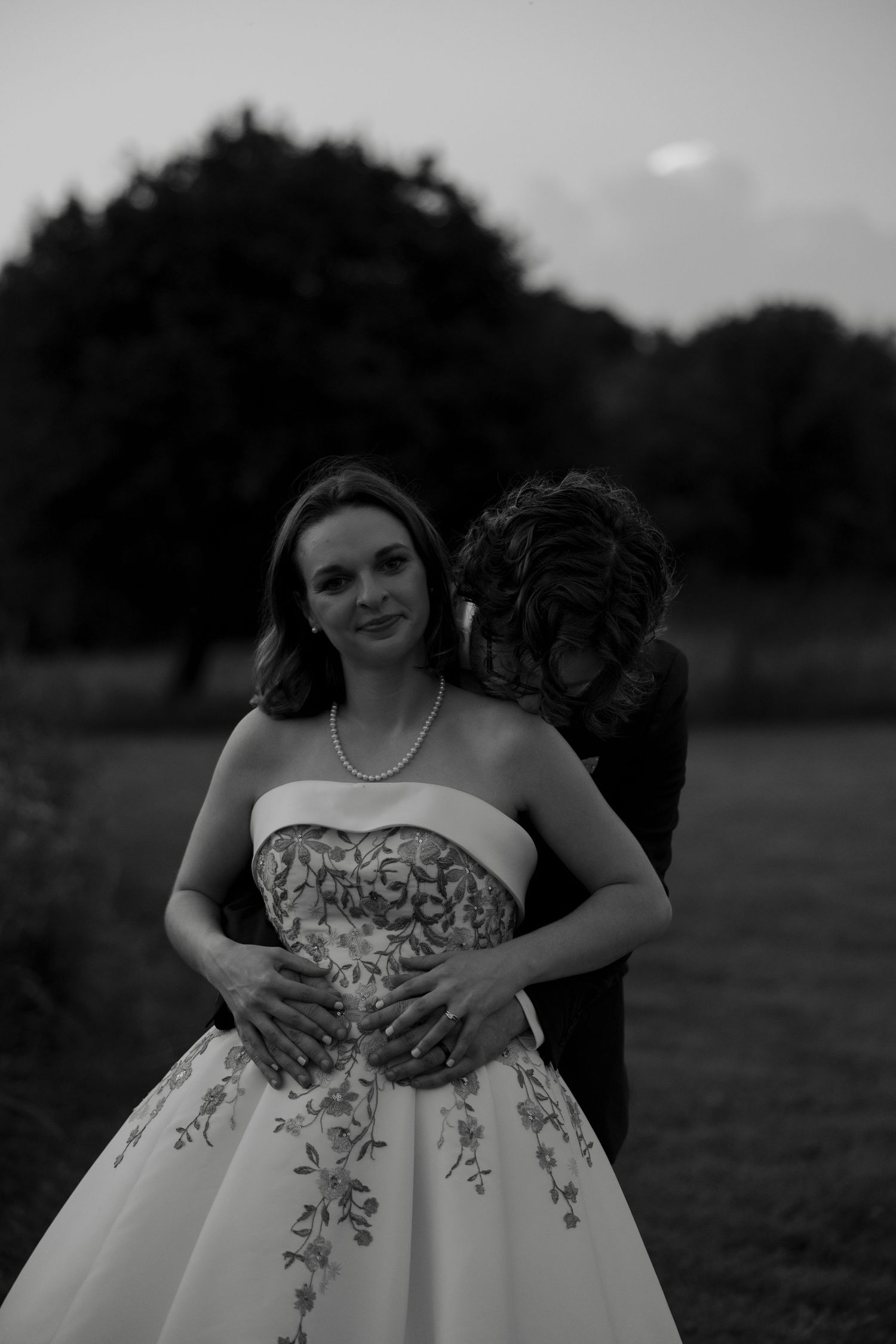 Black and white portrait of bride in strapless floral gown with groom embracing her from behind at dusk.