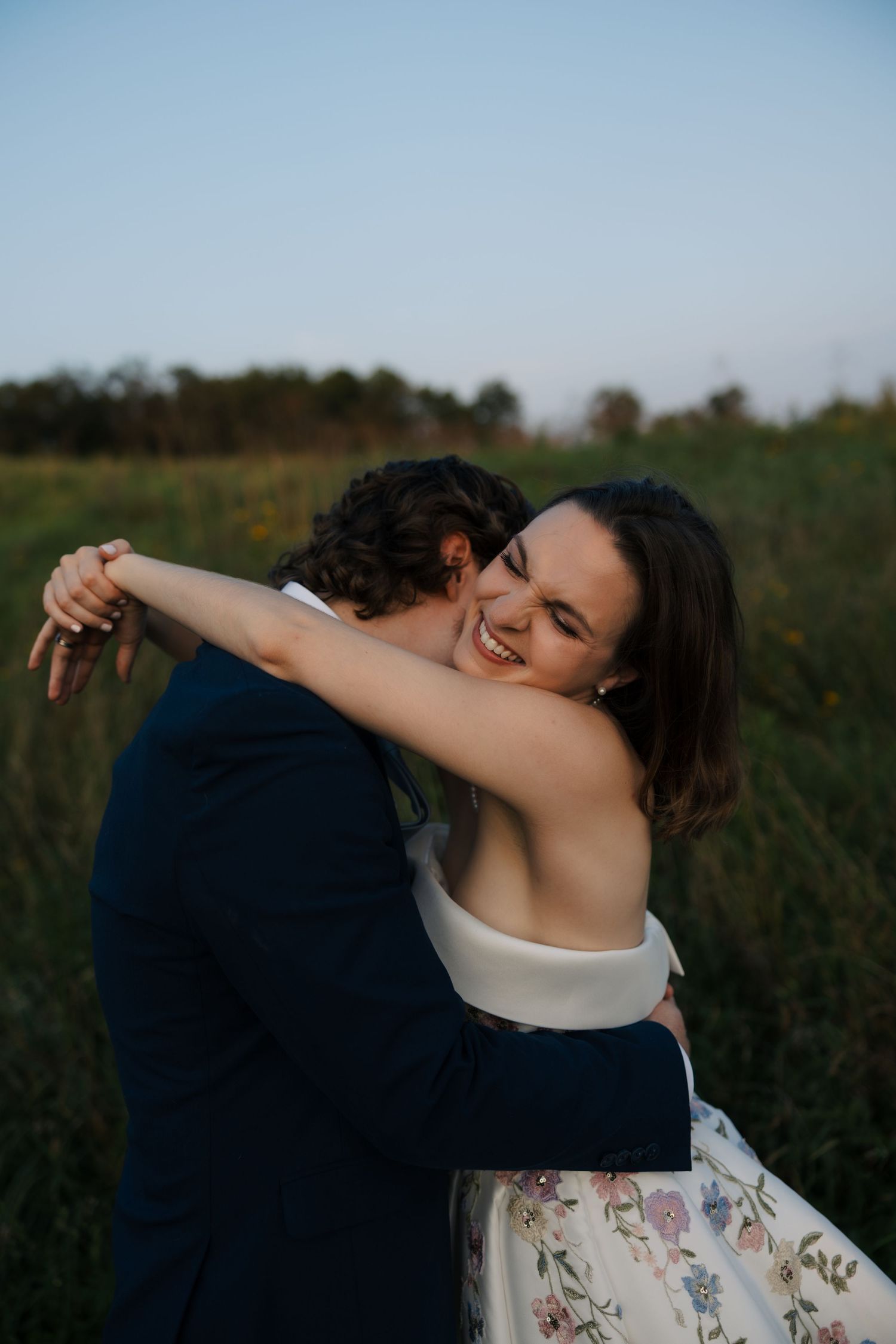 Joyful bride in floral embroidered gown hugs groom tightly while laughing outdoors in a green field at dusk.