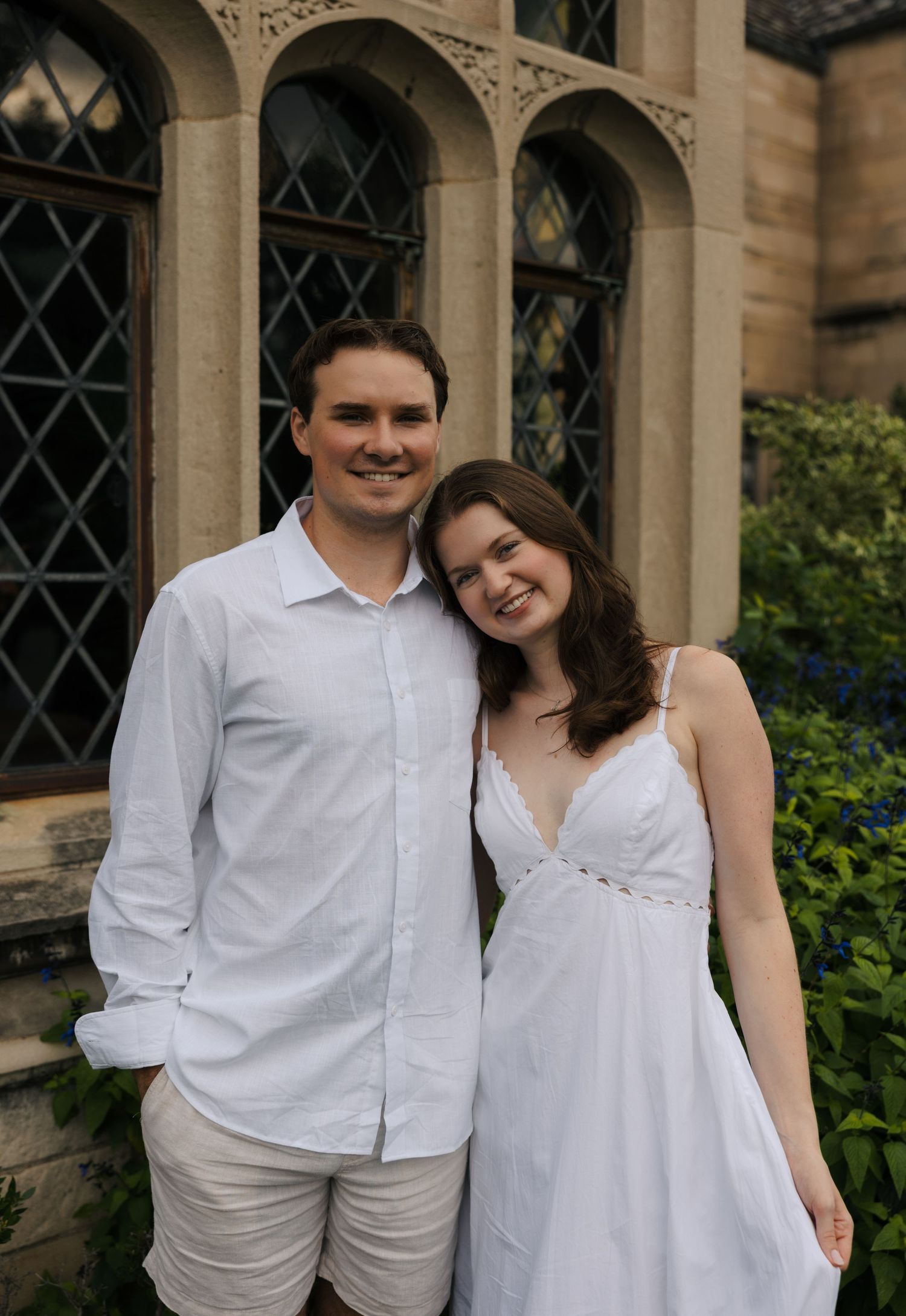 Smiling couple in matching white outfits posing together in front of a stone building with arched windows.