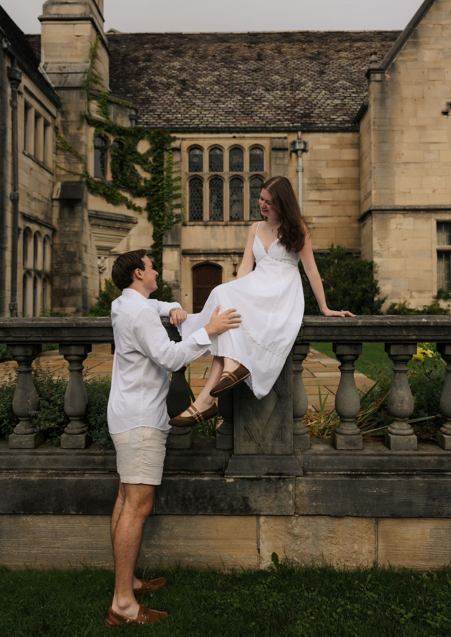 Playful couple in white outfits posing at stone balustrade of a historic manor, woman perched on railing.
