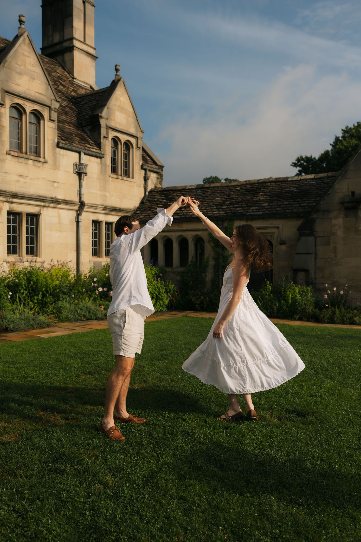 A couple in white outfits dancing on green grass in front of a historic stone manor house on a sunny day.