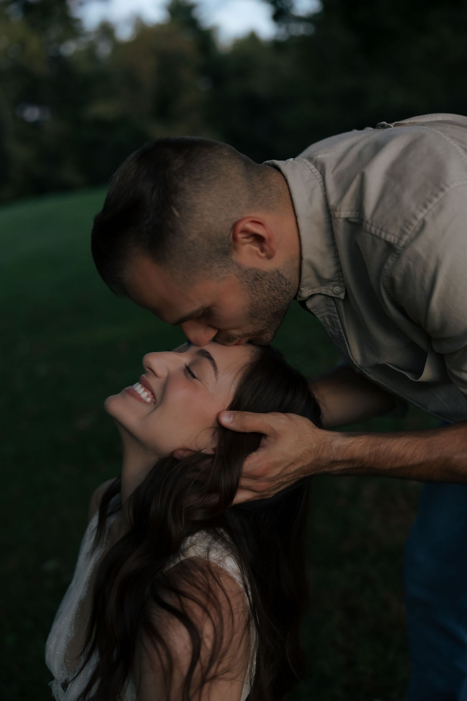 Man kisses laughing woman's forehead while holding her hair in romantic outdoor evening moment.