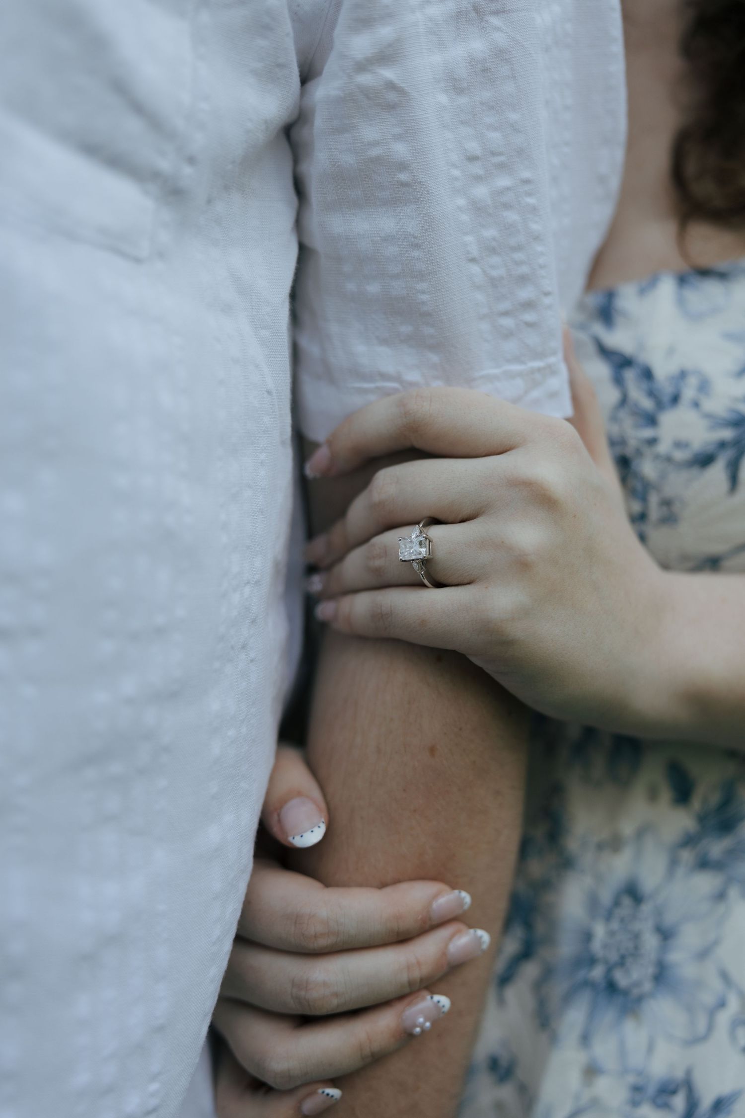 Close-up of a woman's hand with a diamond engagement ring, holding a man's arm, wearing a floral blue dress.