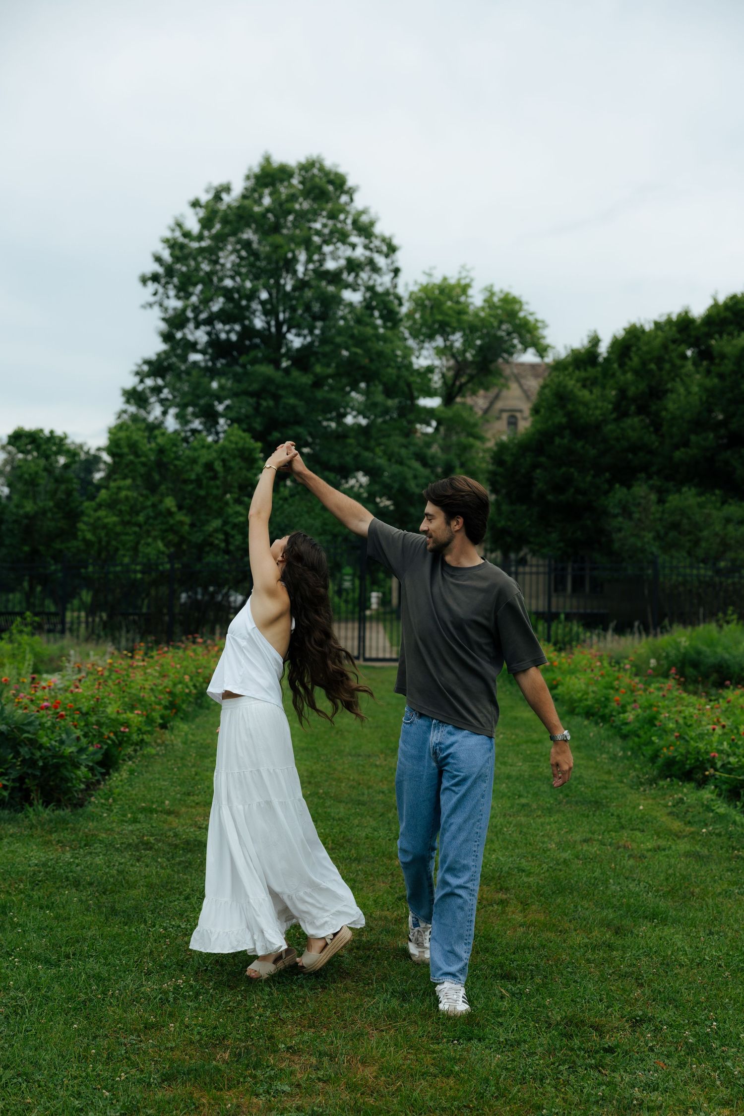 Couple dancing together on lush green grass in a garden, woman in white dress being twirled by man in gray tee.