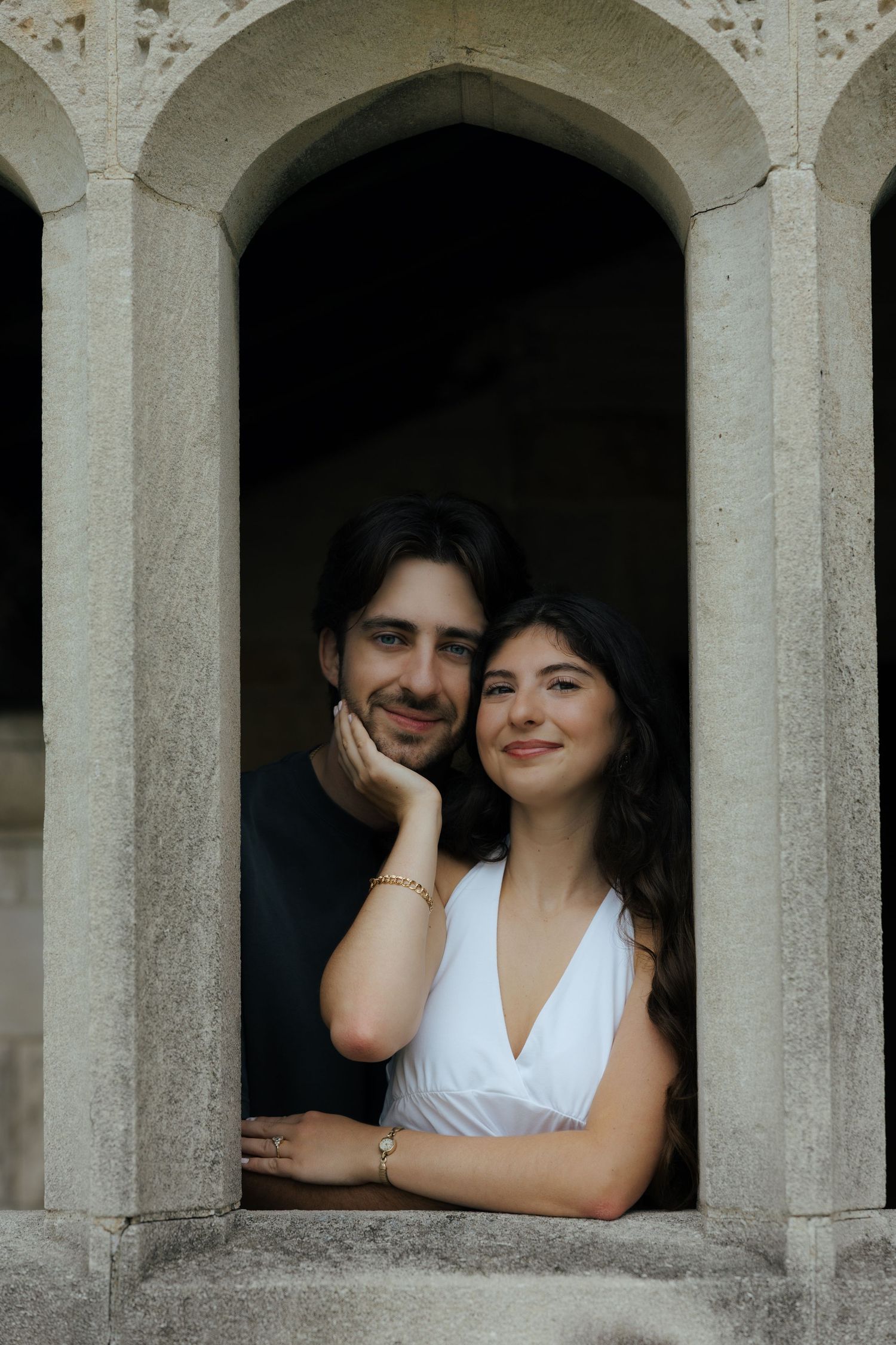 Smiling couple leaning out of a stone arch window, woman in white dress with gold bracelet.