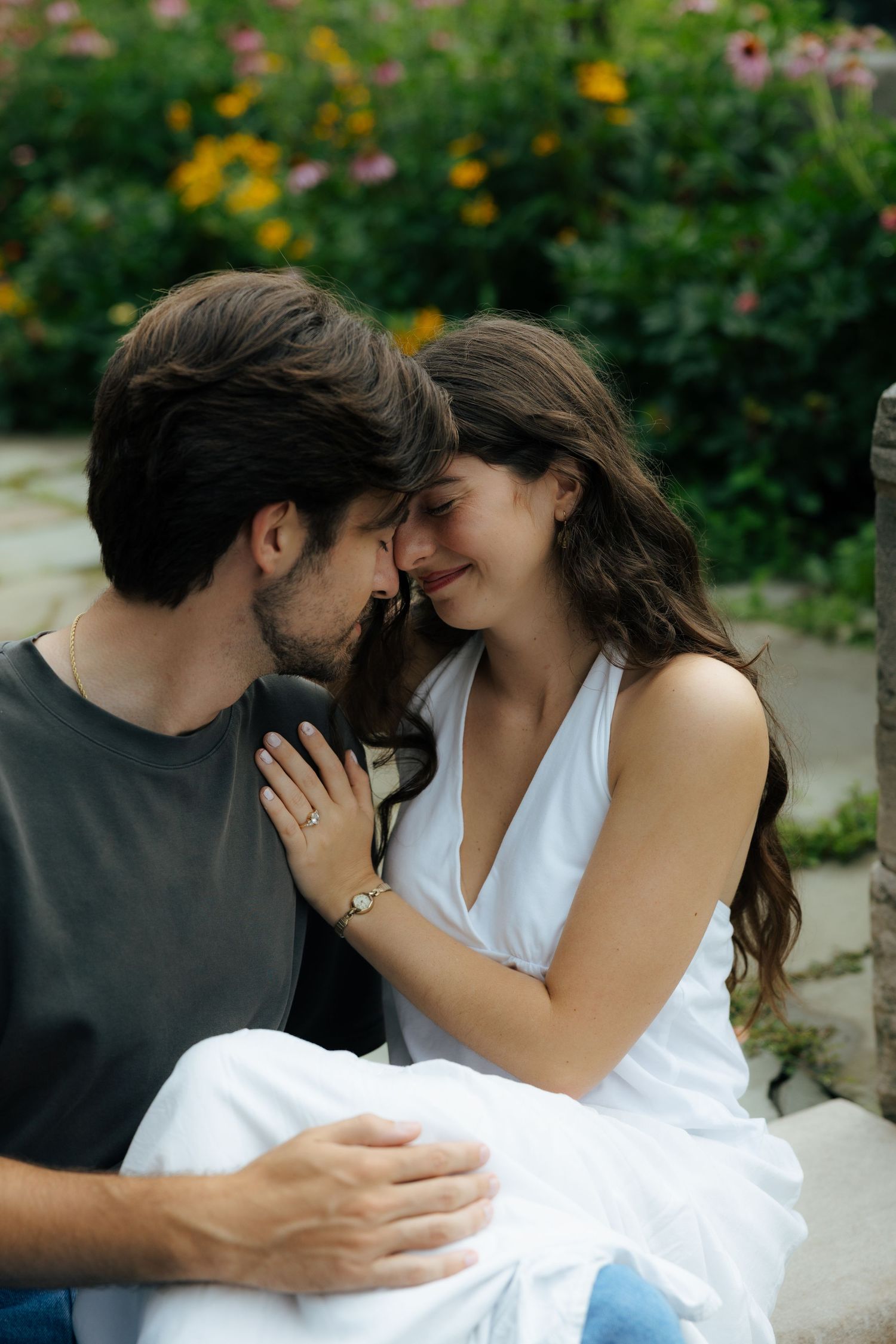 A couple shares an intimate moment, touching foreheads and smiling in a lush garden with colorful flowers.