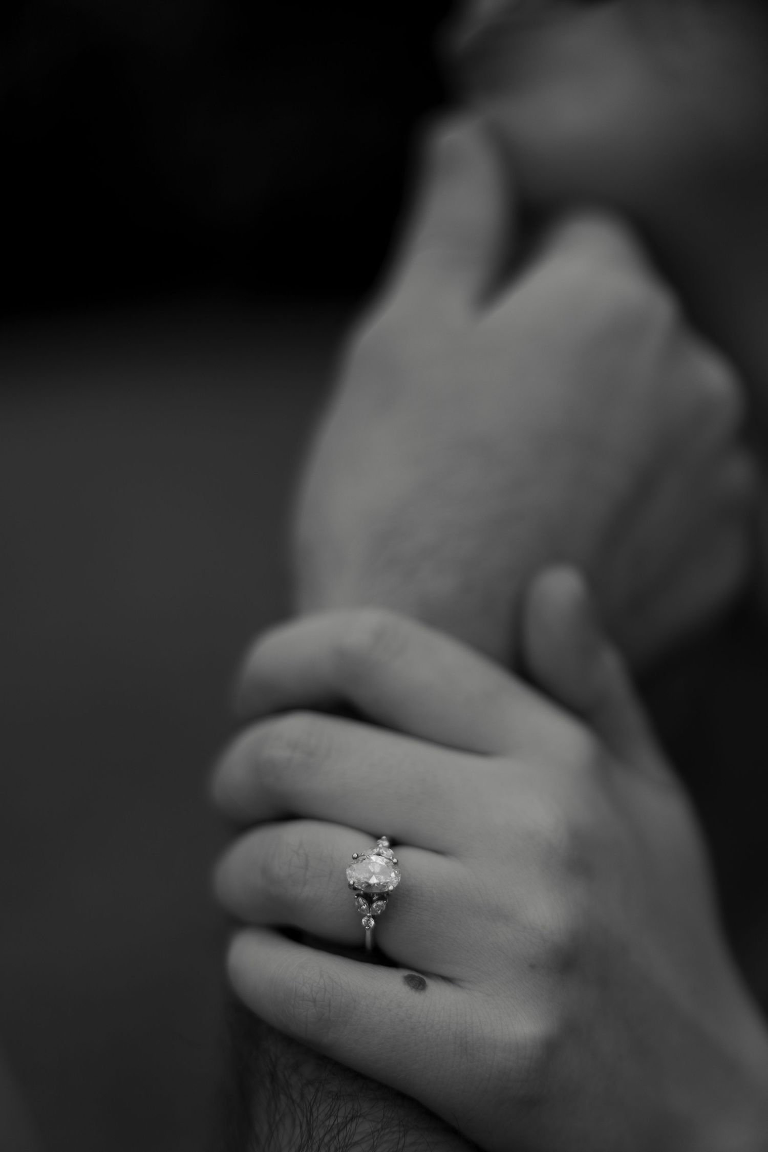 Close-up black and white photo of an elegant diamond engagement ring on a womans hand held by her partner.