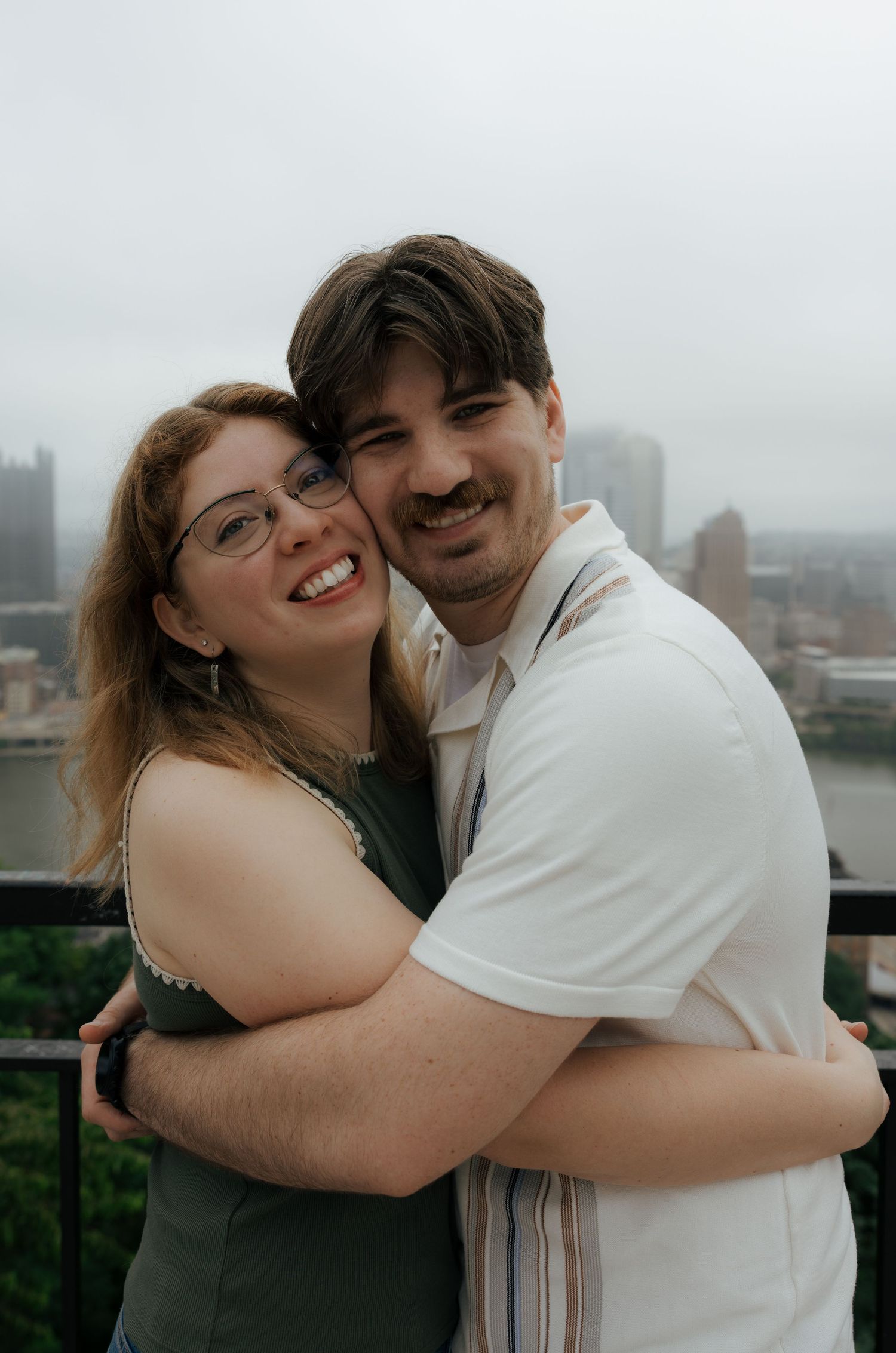 Smiling couple embracing closely on a balcony overlooking a misty Pittsburgh cityscape.