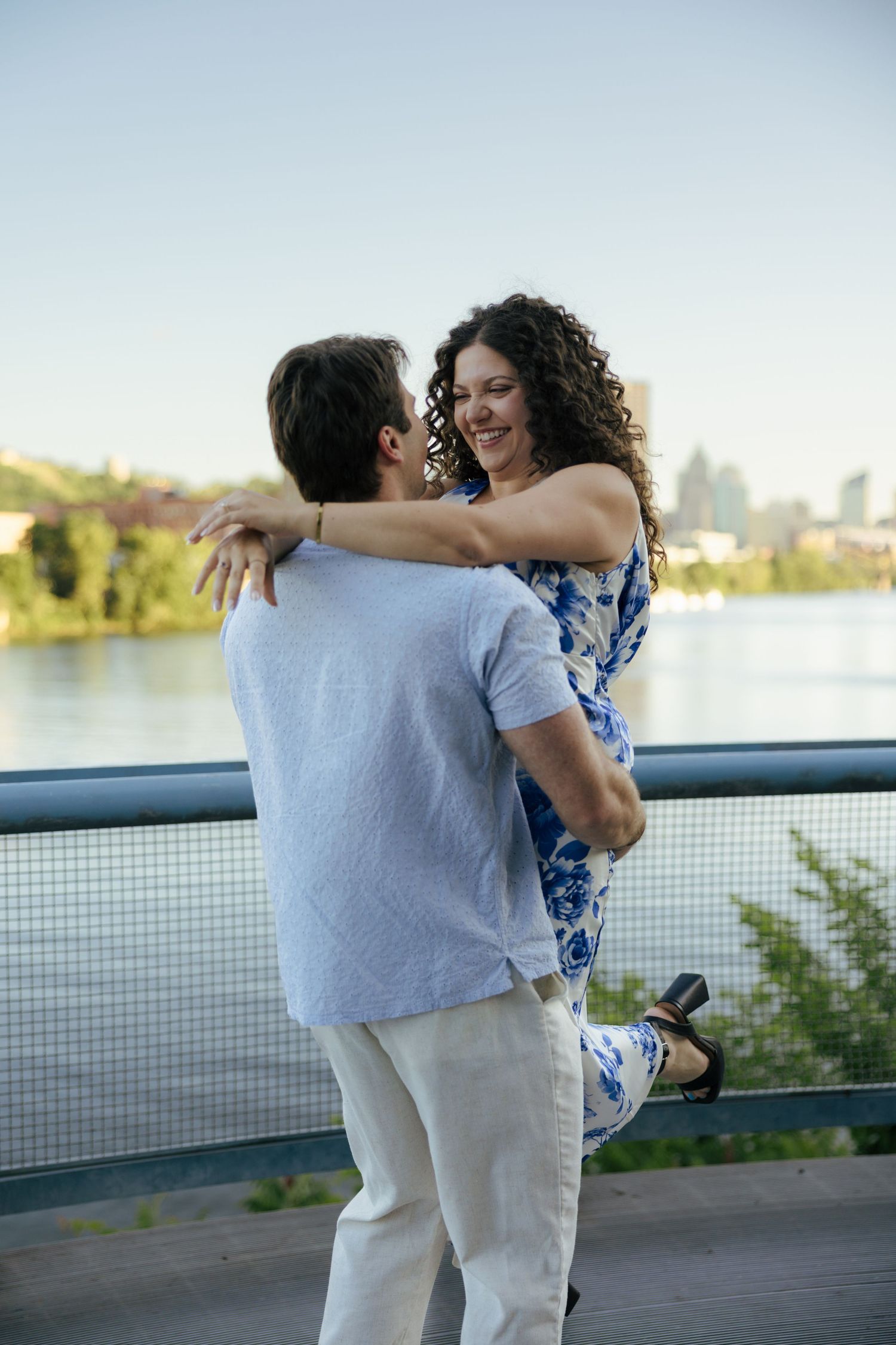 A man lifts a smiling woman in a blue floral dress on a bridge overlooking a sunny river.