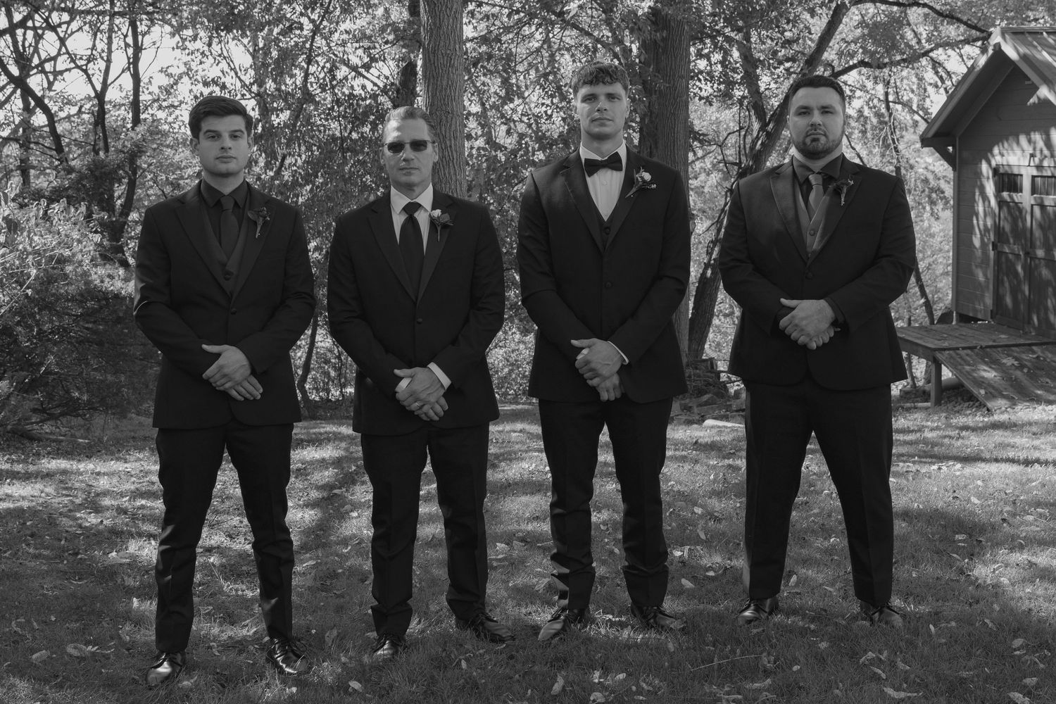Four men in black suits posing outdoors for a formal group photo in a black and white setting.